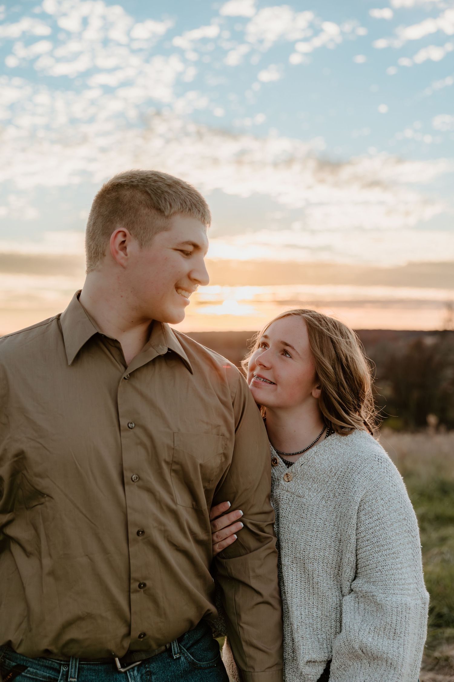 A couple shares a tender moment at sunset while wearing a brown shirt and gray sweater in an outdoor setting.