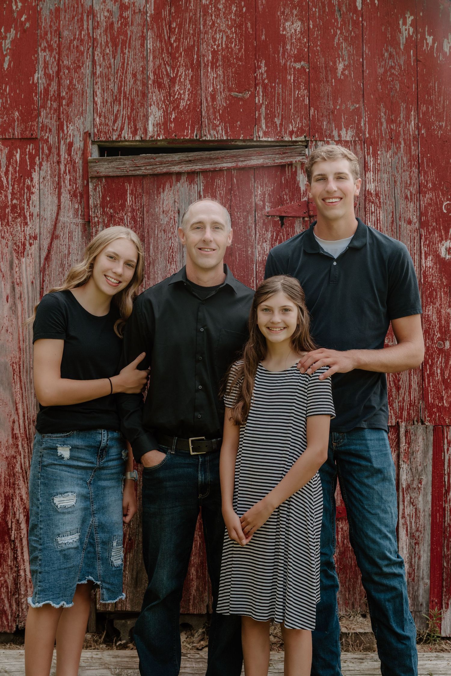 Family group wearing black and denim poses in front of a rustic red barn wall for a casual portrait session.