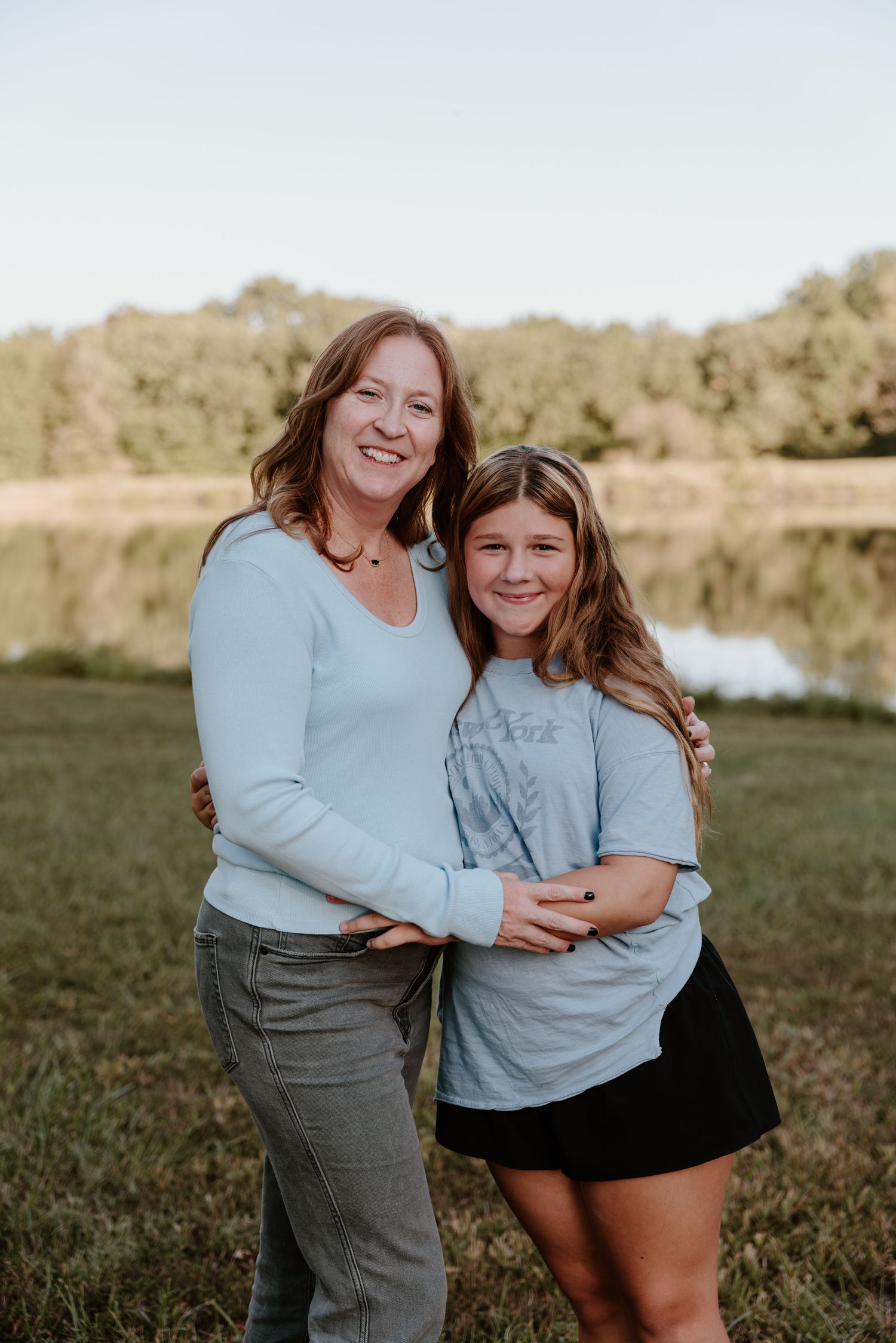 Two people in matching light blue tops share joyful embraces outdoors by a pond on a sunny afternoon.