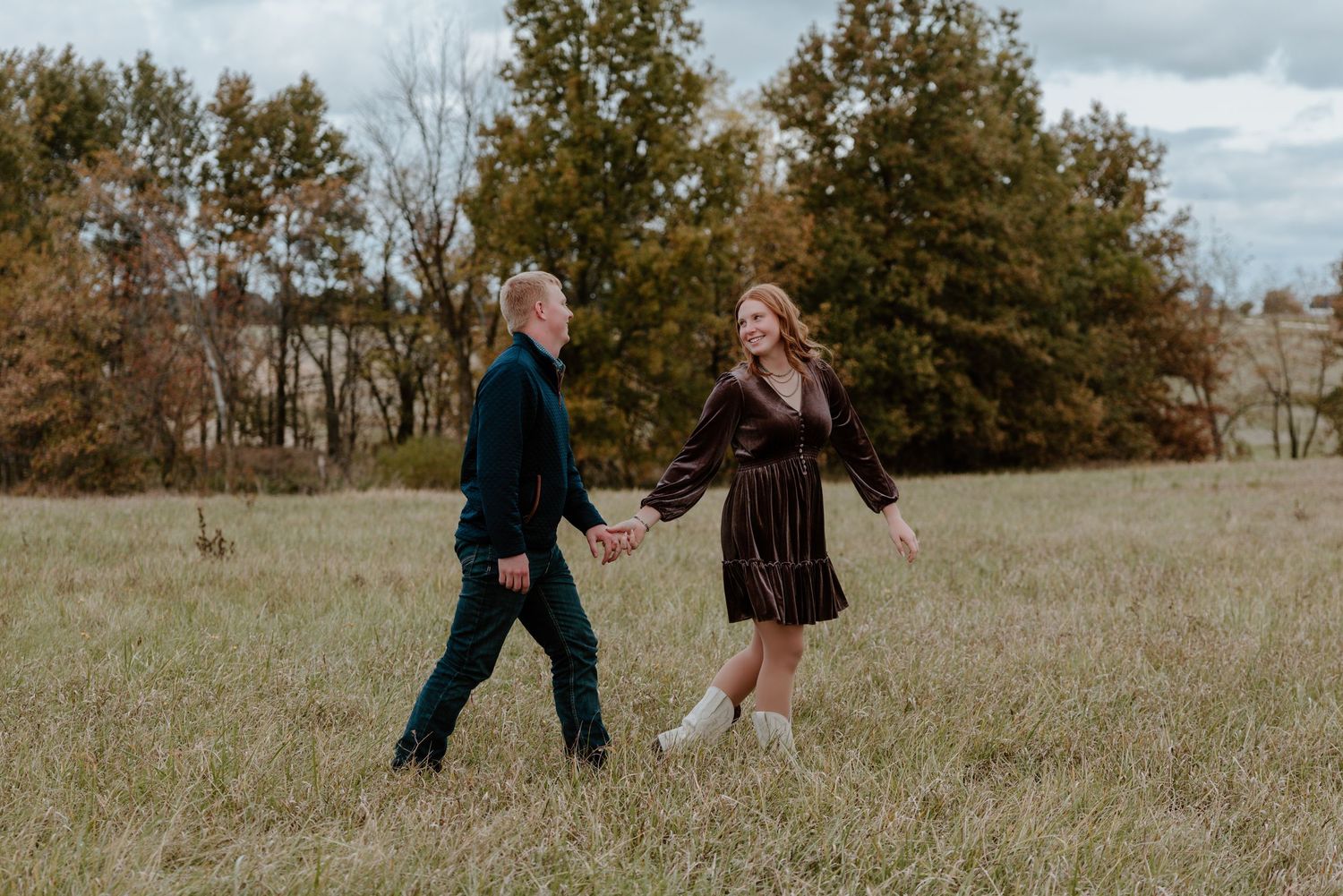 Two people hold hands while walking through a golden autumn field on a cloudy day.