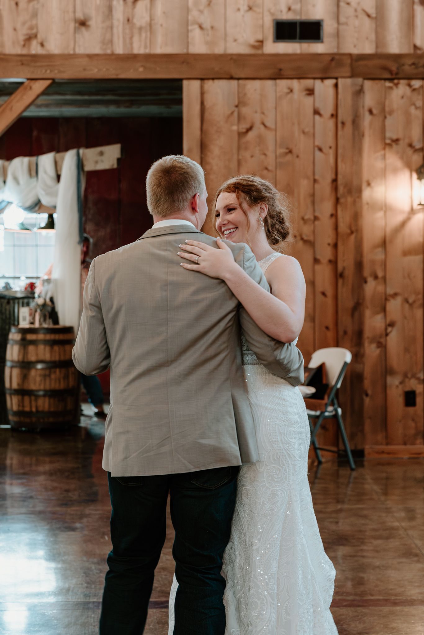 Bride and groom share their first dance in a rustic barn wedding venue, bride smiling joyfully in a lace gown.