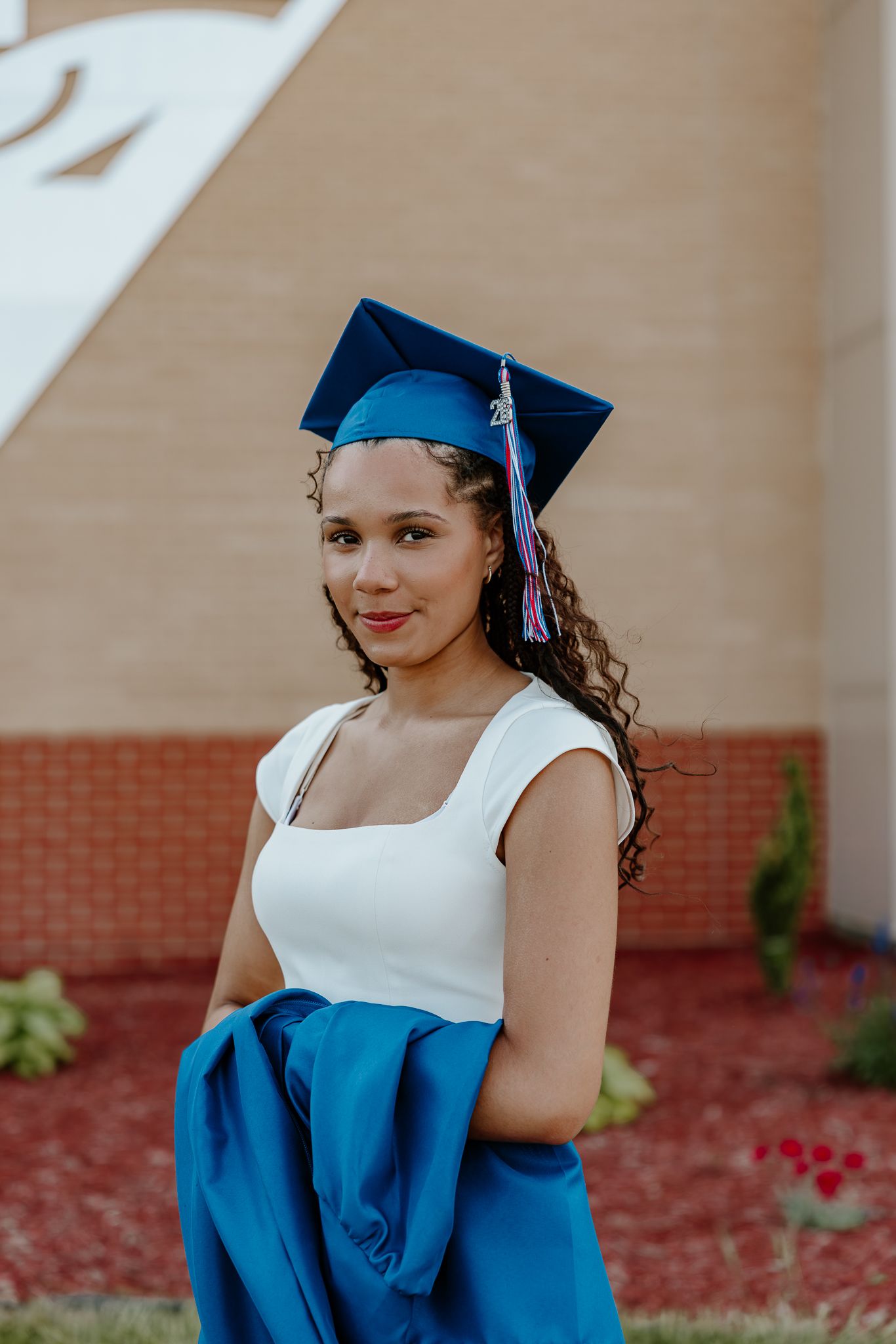 Young woman in blue graduation cap and gown smiles outdoors near brick building on graduation day.