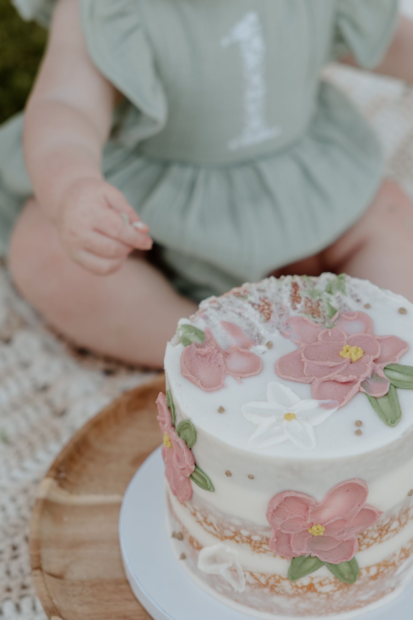 A floral first birthday smash cake with pink flowers sits in front of a baby wearing a green romper with number 1.