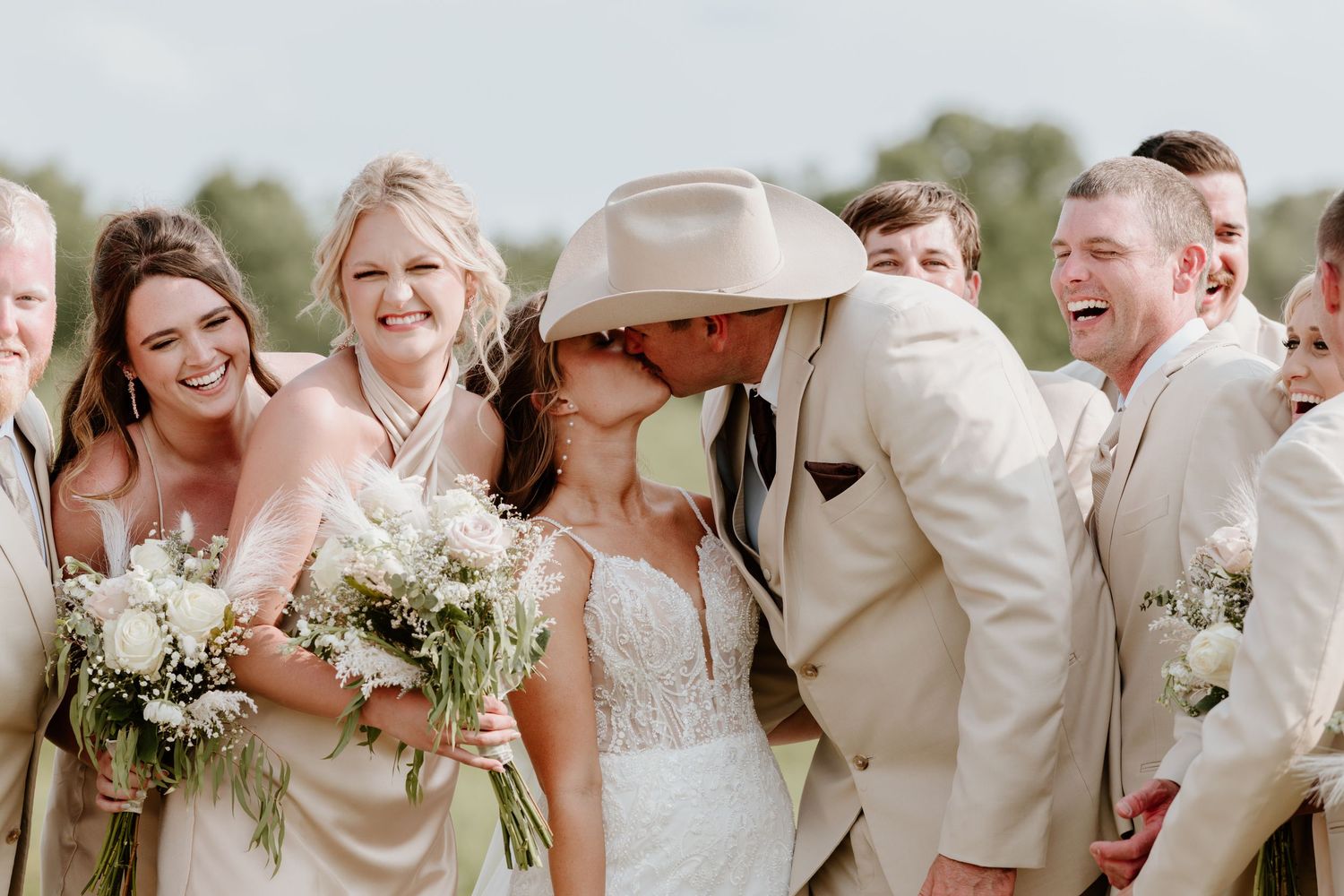 Bride and groom sharing a kiss surrounded by laughing wedding party in champagne attire.