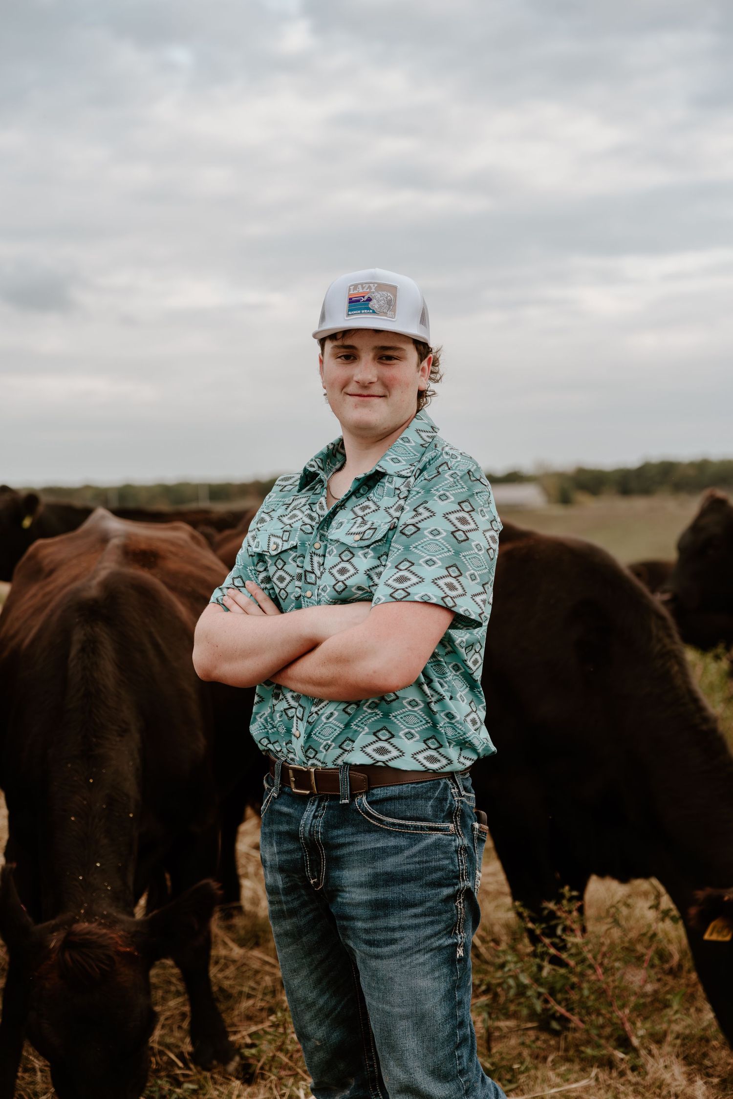 A person in a patterned shirt and baseball cap stands confidently in front of grazing cattle on a ranch.