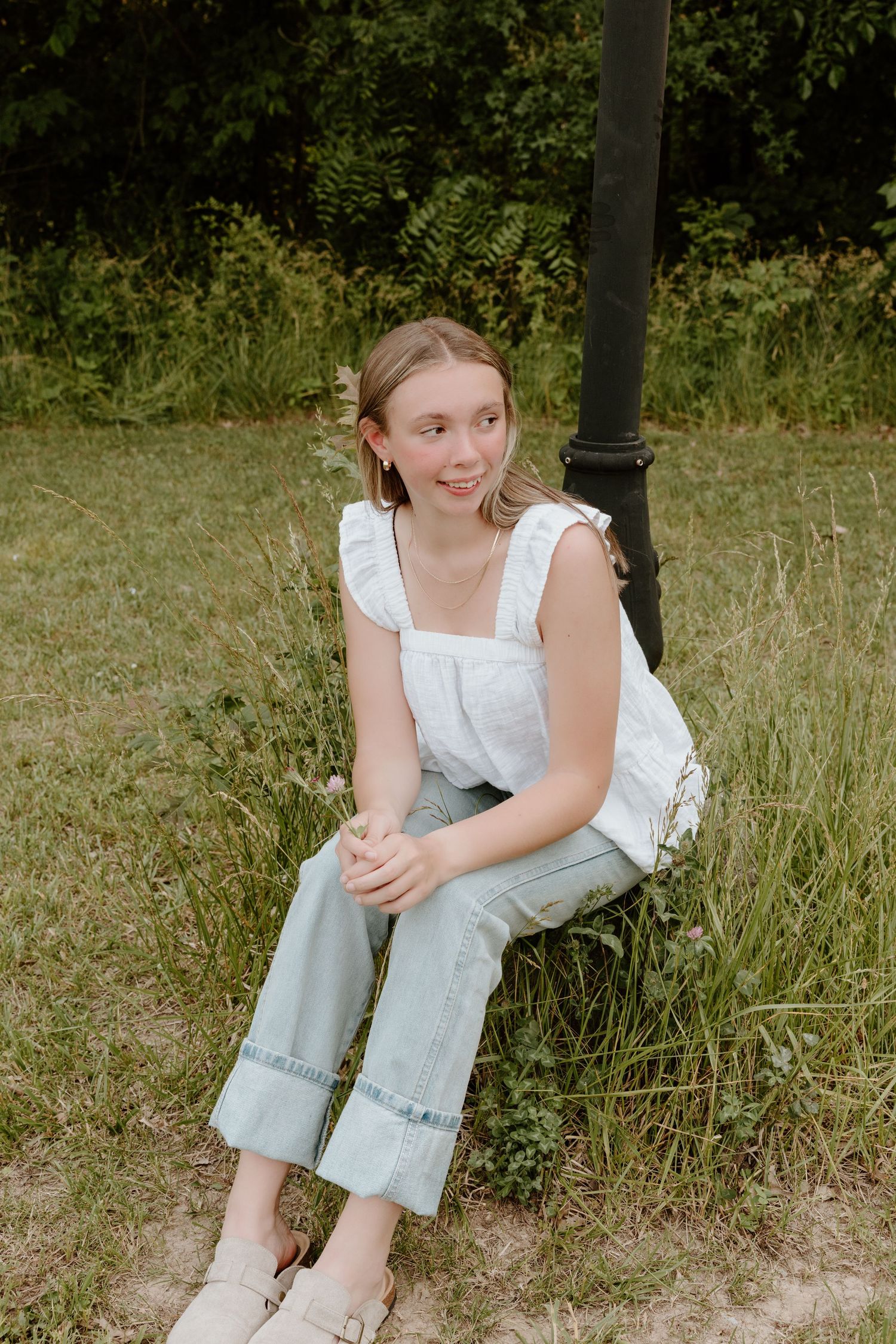 Someone sits casually outdoors wearing white overalls and a ruffled top against a natural green background.