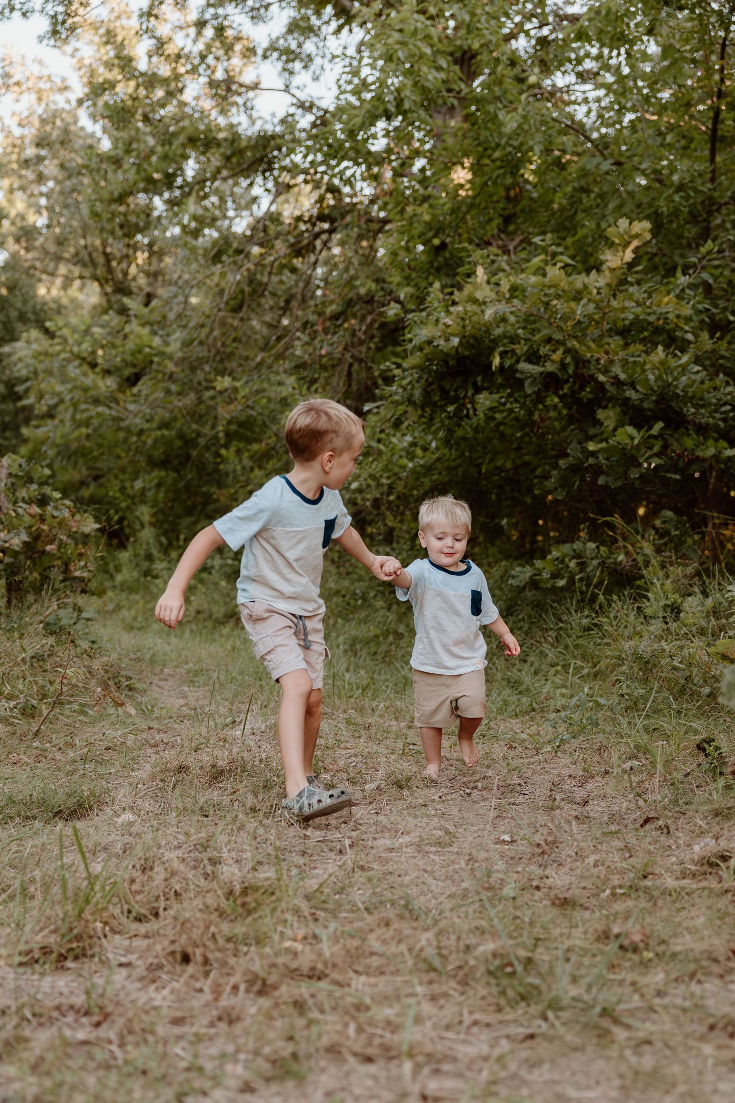 Two children in matching outfits run and play together on a dirt path surrounded by green trees in natural sunlight.