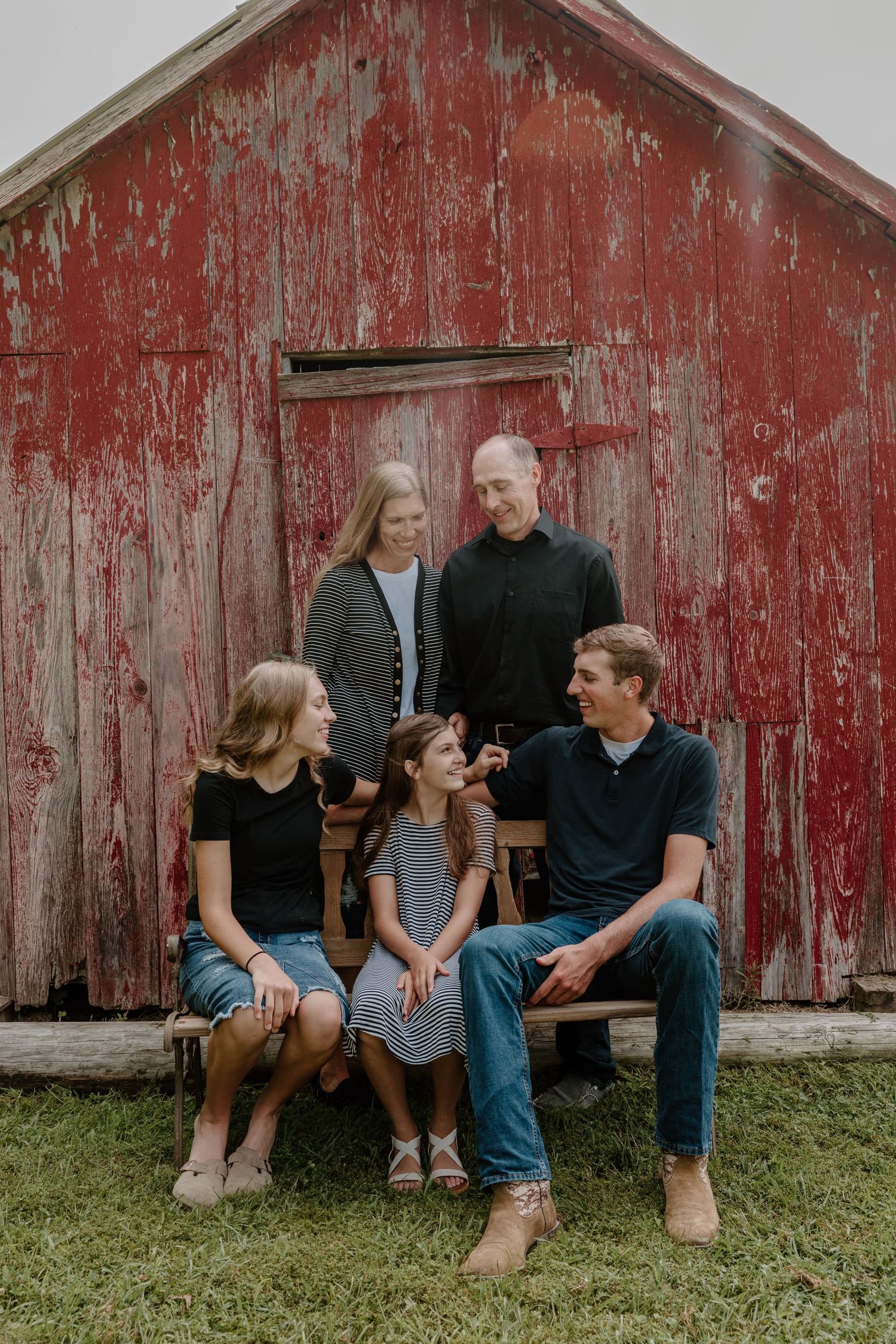 A family sits together and laughs while leaning against a weathered red barn wall during an outdoor photo session.