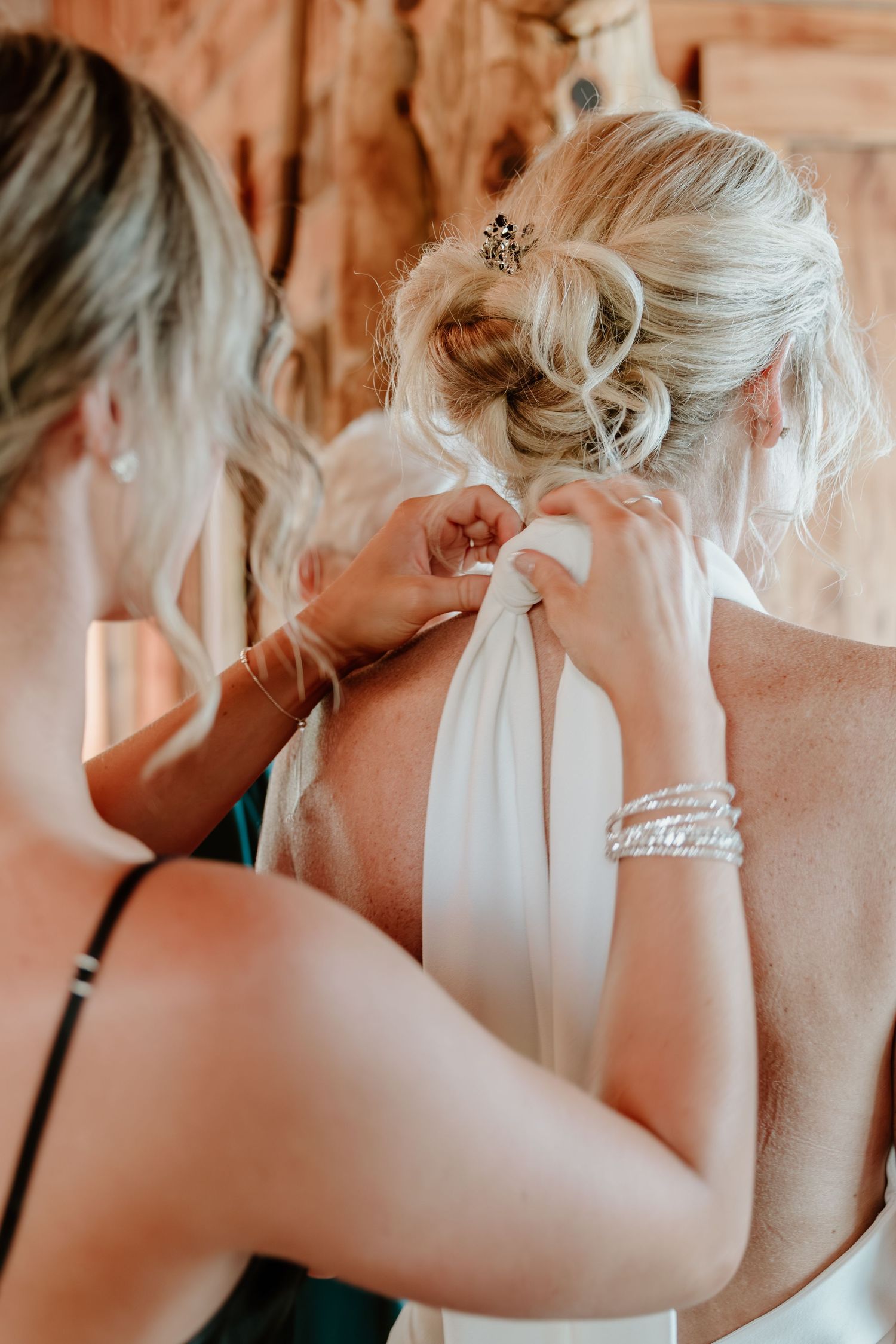 Bridesmaids help tie the back of bride's halter wedding dress with elegant updo and hair accessory.