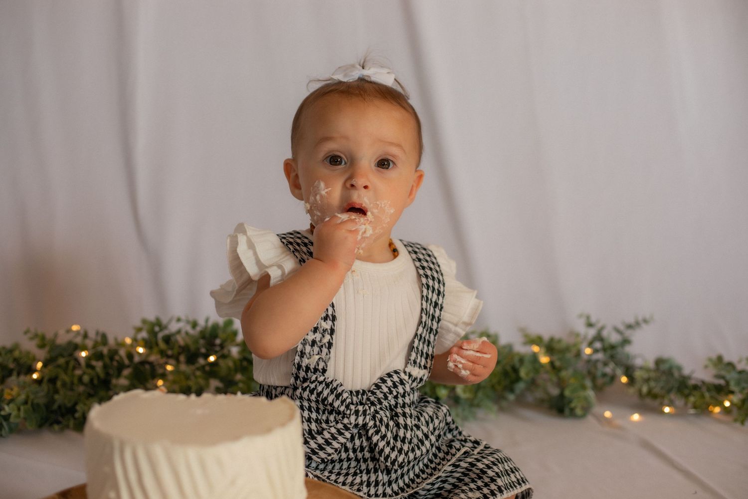 Baby in checkered outfit reaches for white birthday cake with twinkle lights in background.