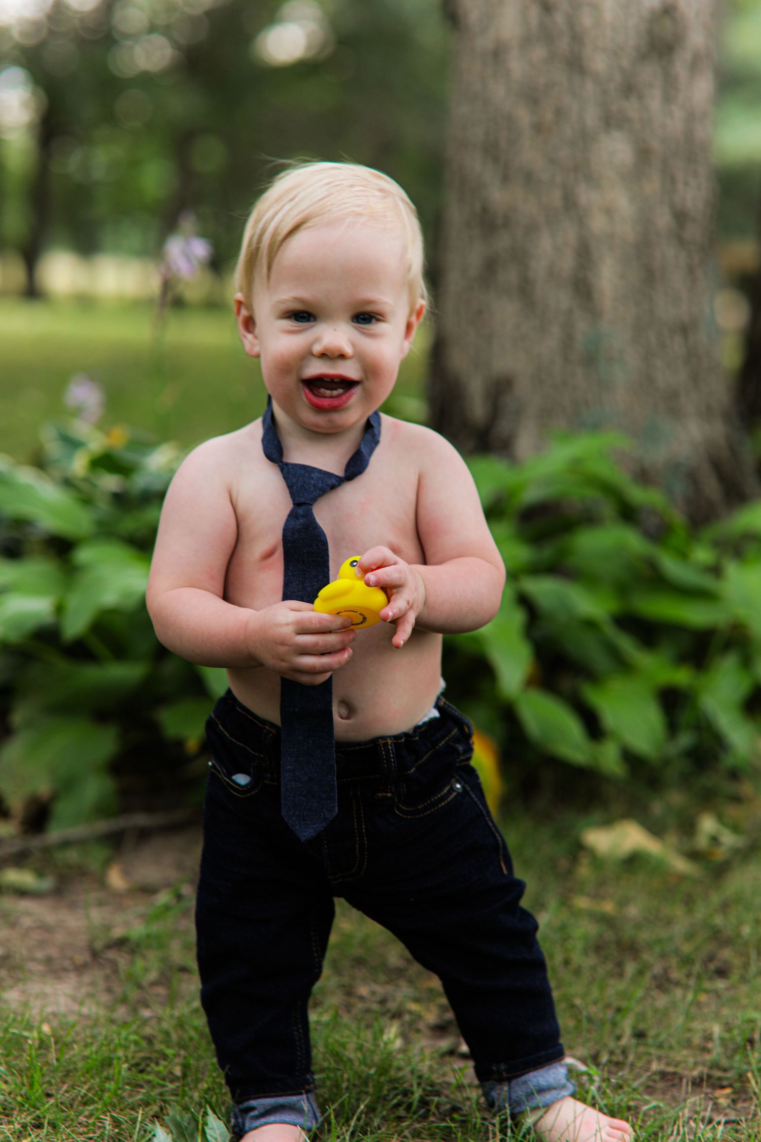 Happy toddler in jeans and navy necktie holds yellow rubber duck outdoors.