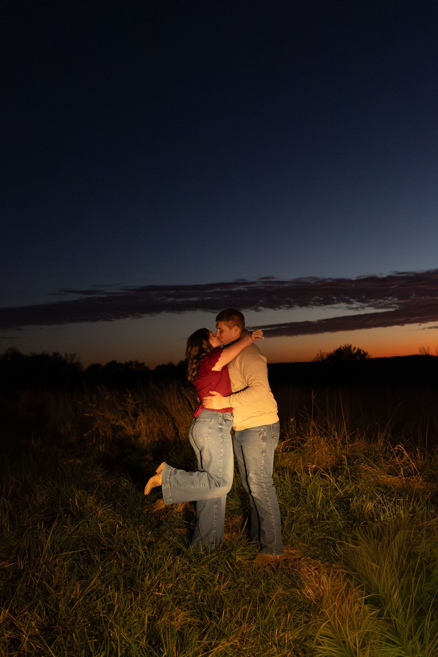 Two people embrace while watching a colorful sunset over distant mountains and grasslands.
