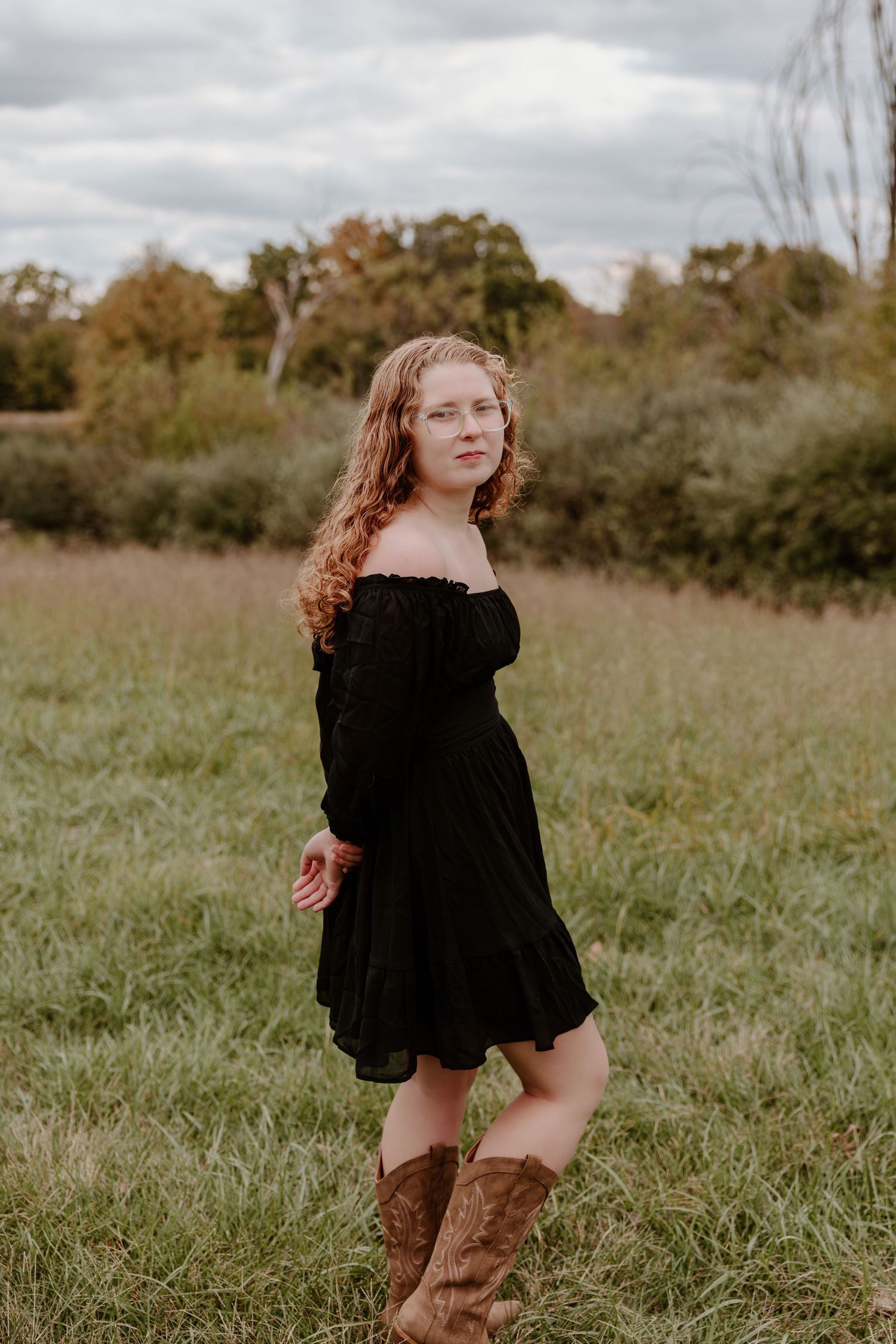 A person in a black dress and brown boots twirls playfully in a grassy field on an overcast day.