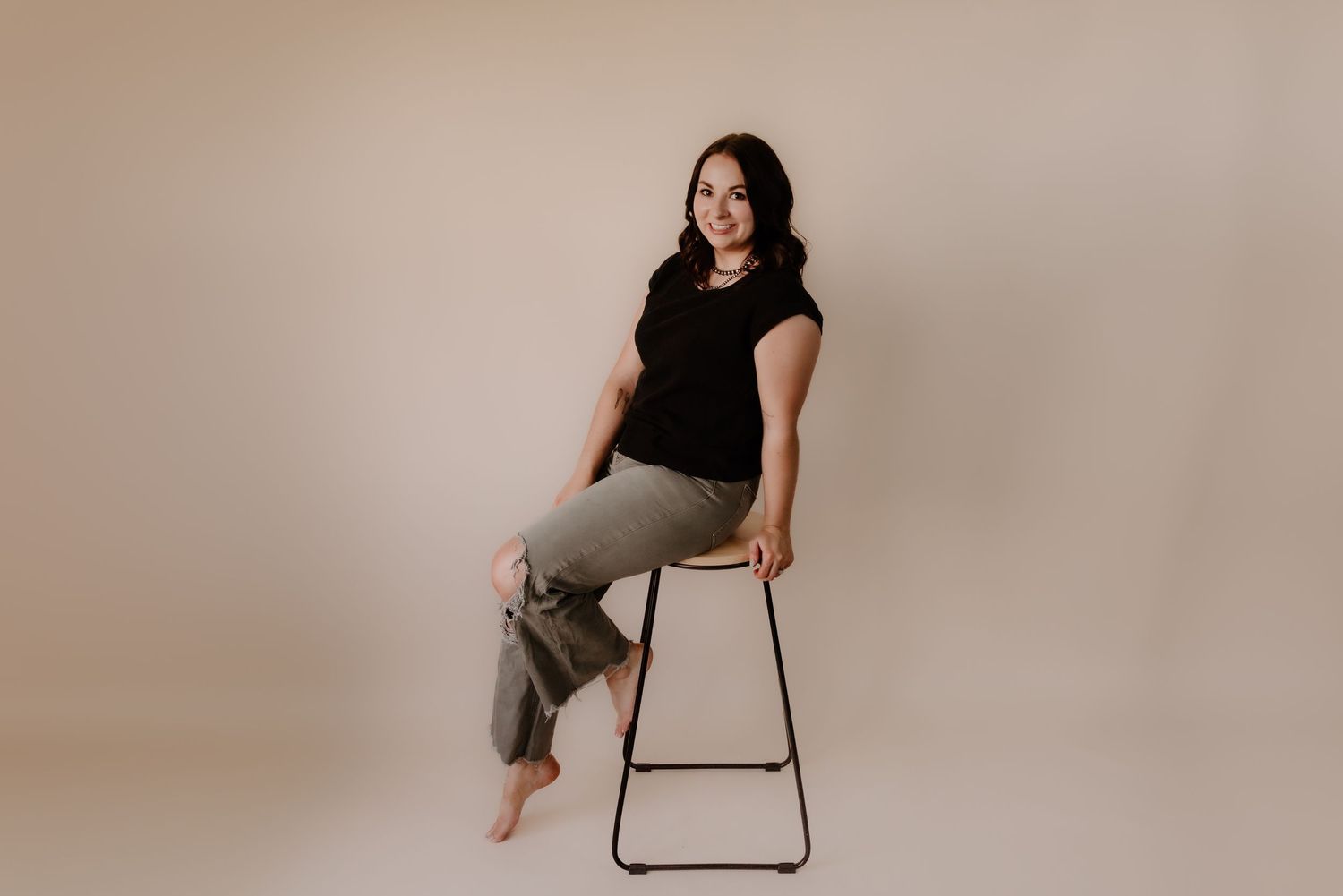 A person in a black top and khaki pants sits casually on a tall stool against a neutral beige studio backdrop.