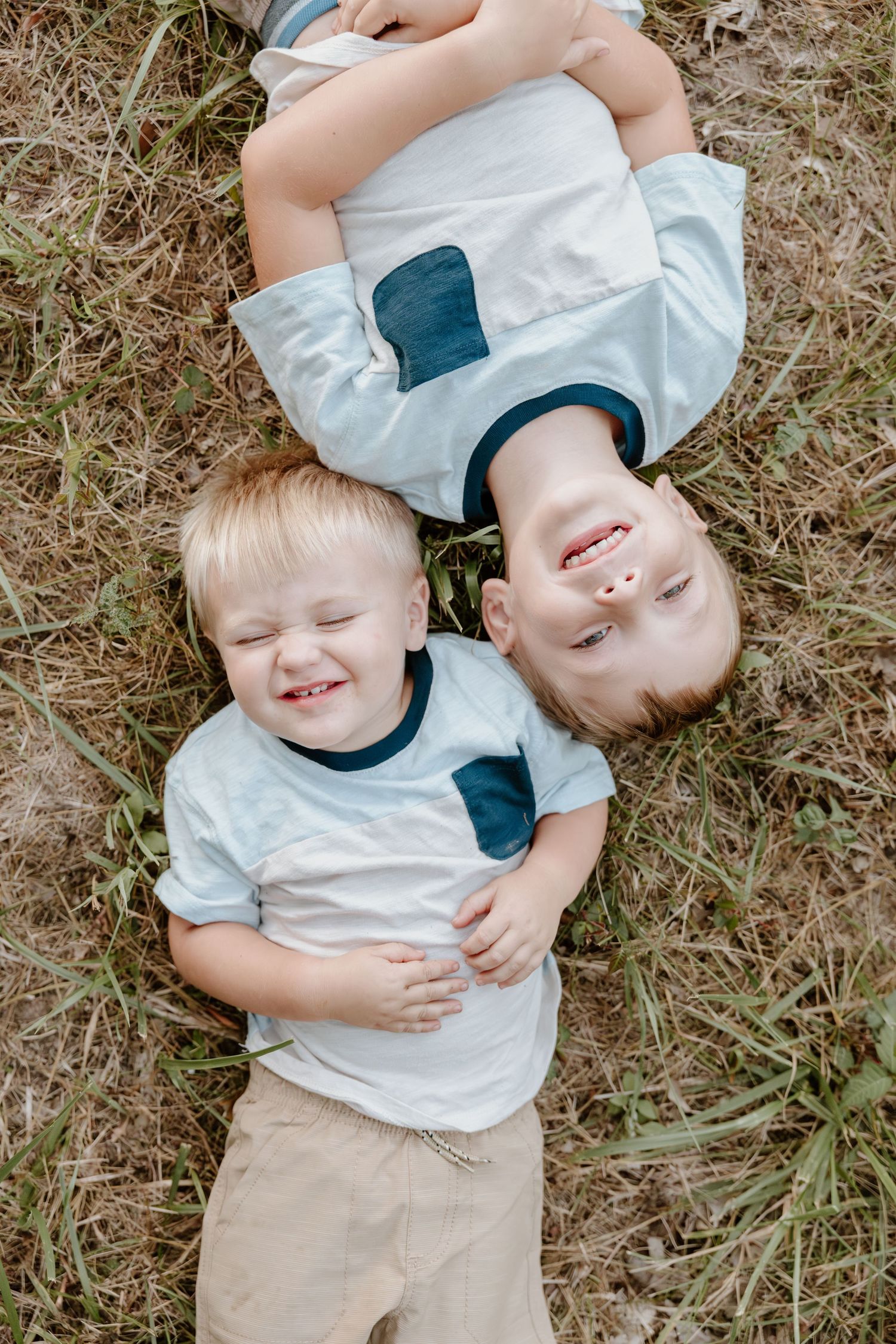Two young children lying on grass wearing matching white shirts and beige pants laugh together during outdoor photoshoot.