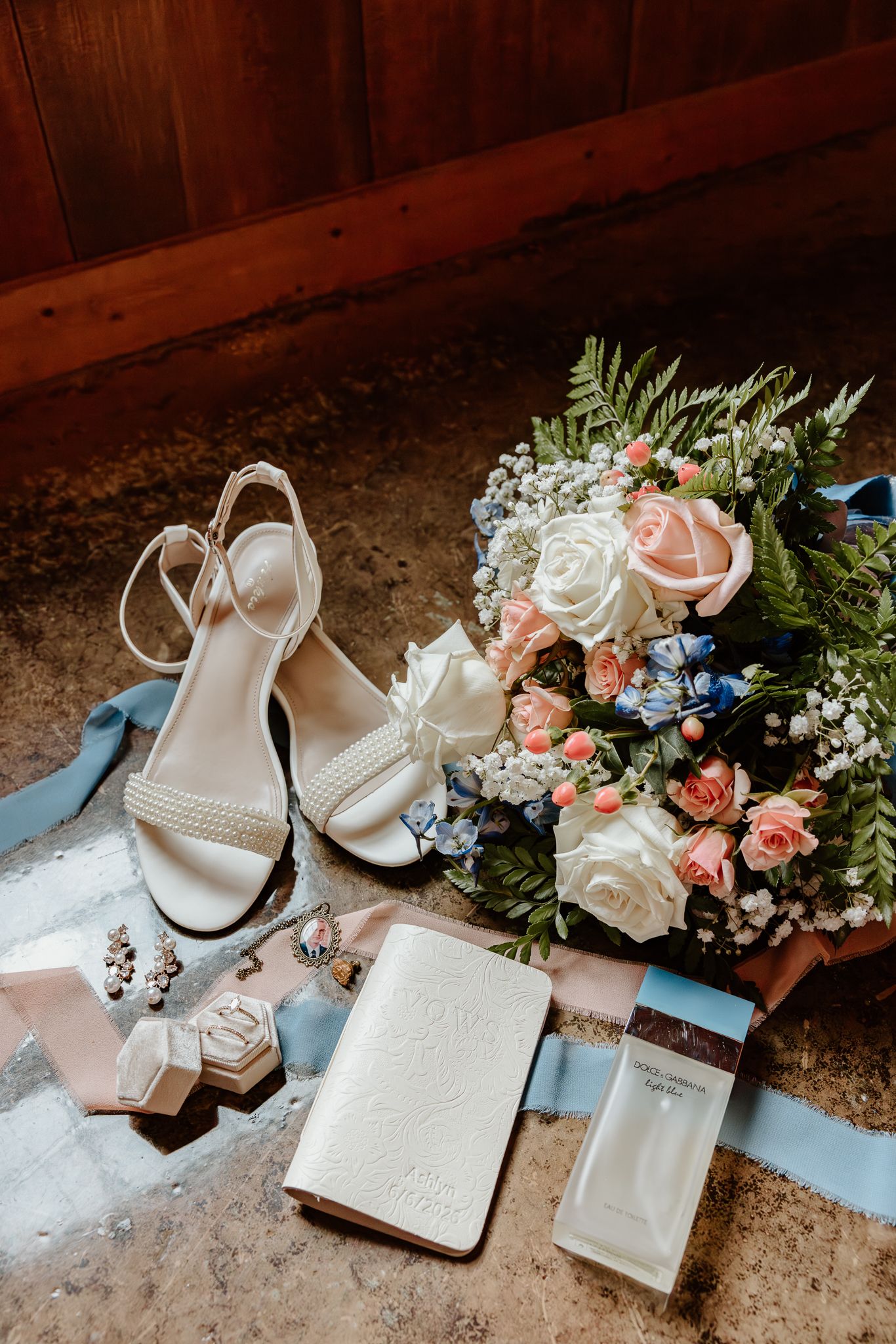 Bridal flat lay with pearl heels, floral bouquet, ring box, veil, and perfume on marble surface.