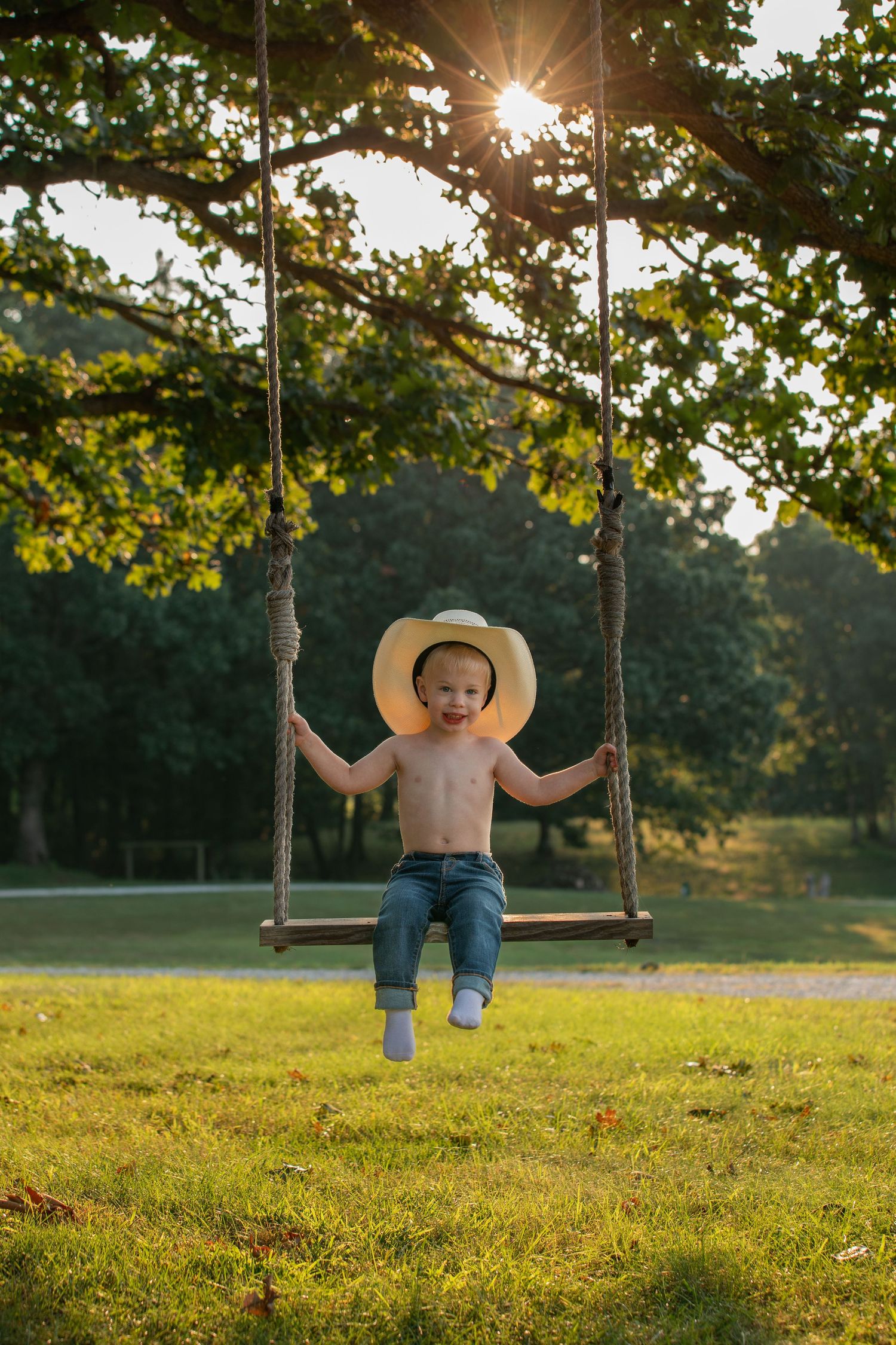 Child wearing cowboy hat swings on wooden swing during golden sunset.