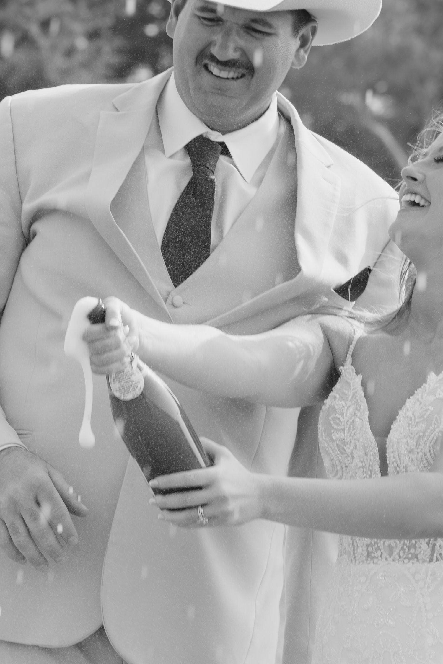A laughing couple pops a champagne bottle at their wedding, with the groom wearing a white suit and cowboy hat.