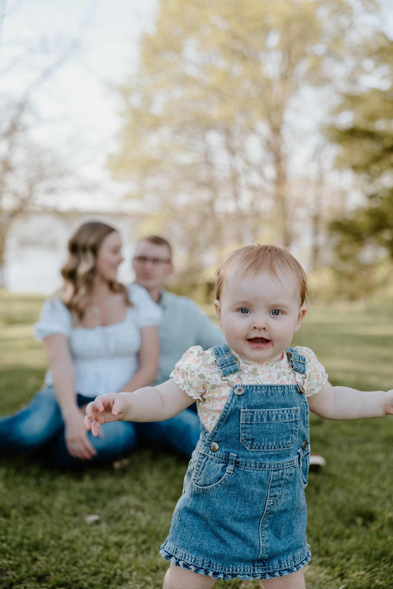 Smiling baby girl in denim overalls taking first steps outdoors while parents sit on grass behind her.