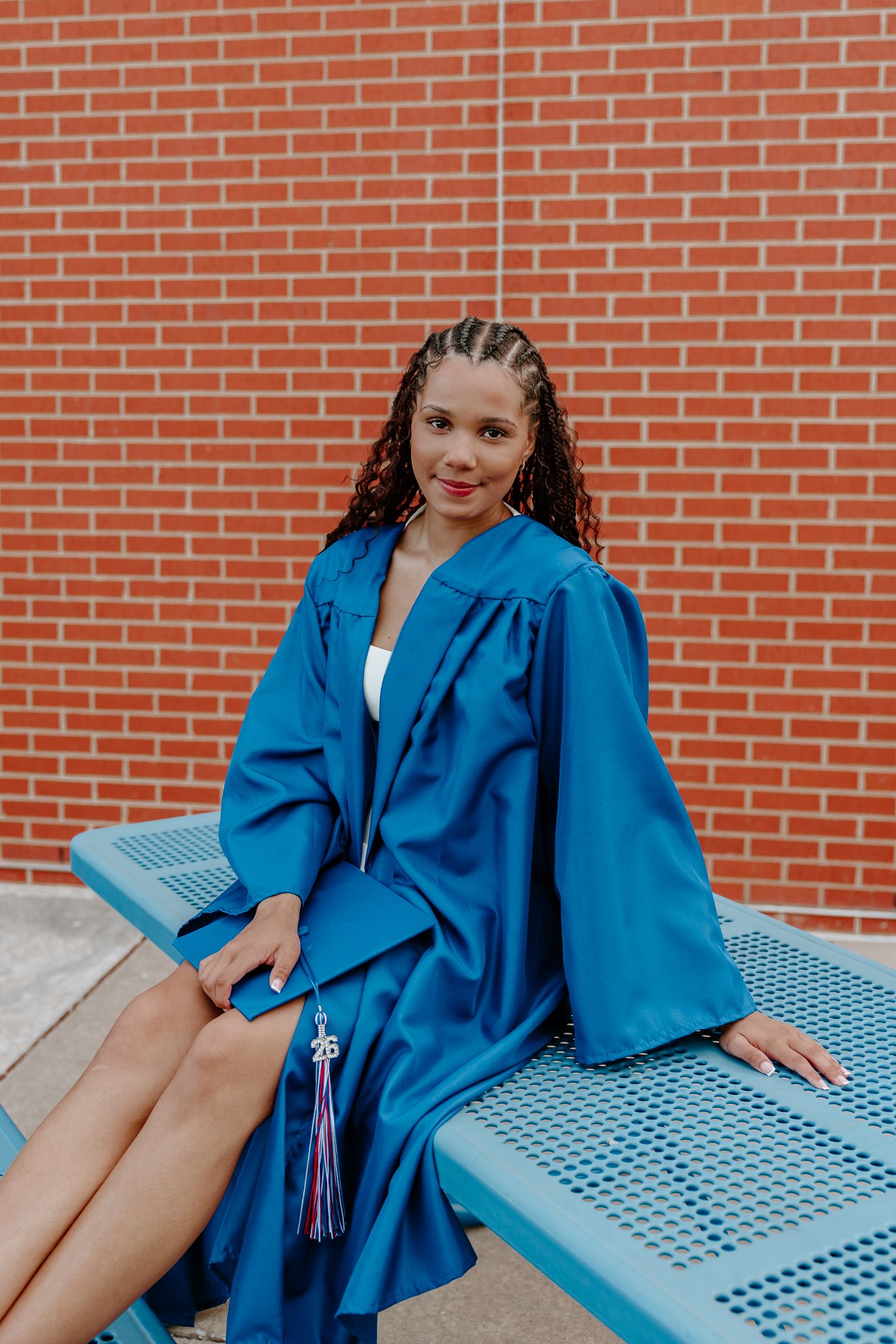 Young woman in blue graduation cap and gown sitting on bench against red brick wall, smiling for senior portrait.
