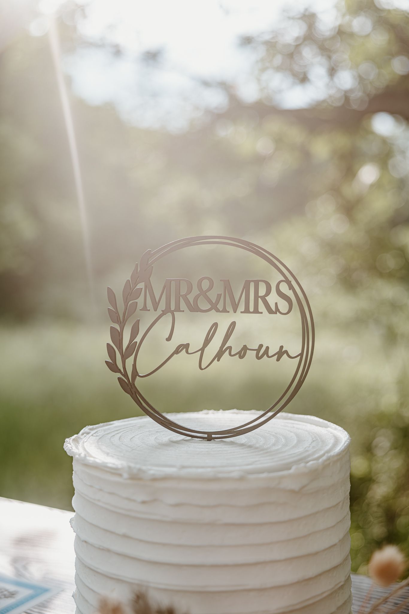 White wedding cake topped with a wooden Mr & Mrs Calhoun circular cake topper, set outdoors with greenery background.