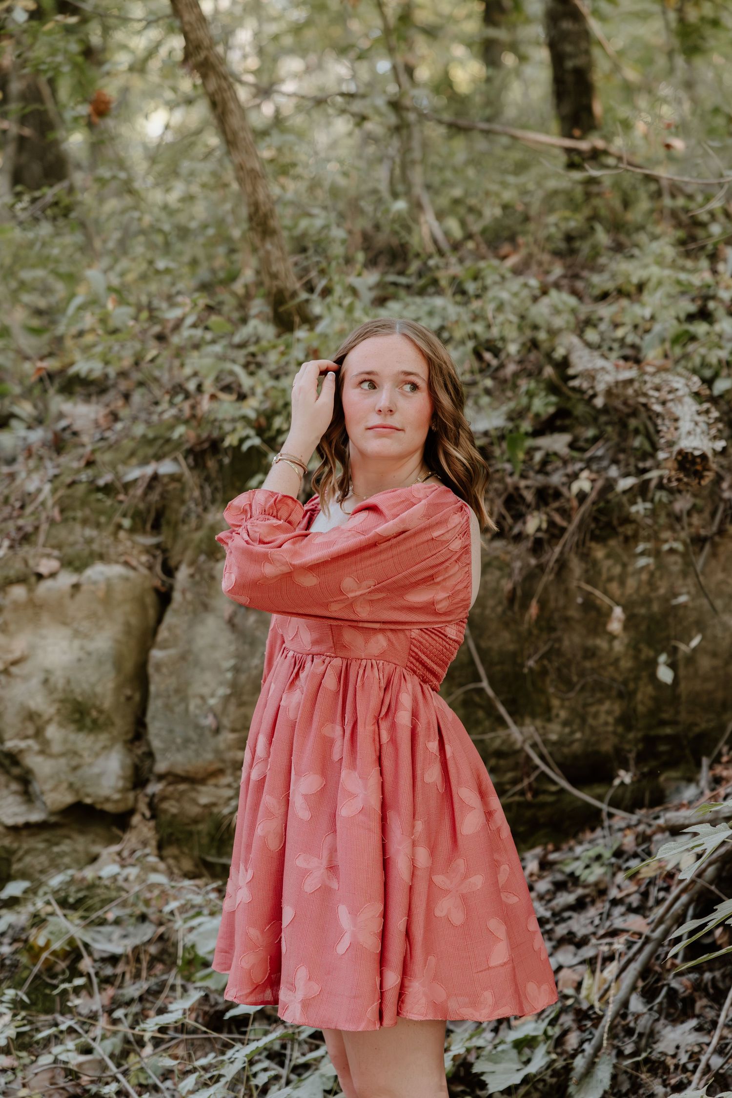 A person in a coral pink dress poses thoughtfully against a natural stone backdrop in a forest setting.