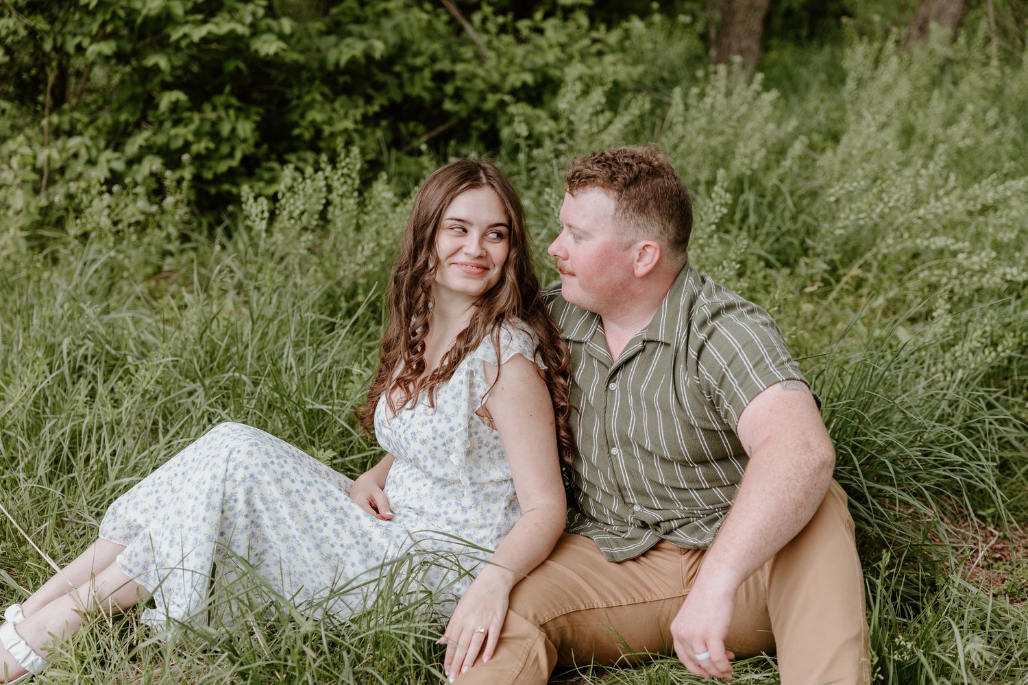 A smiling couple sits together in a lush green meadow, the woman in a floral dress looking at her partner.
