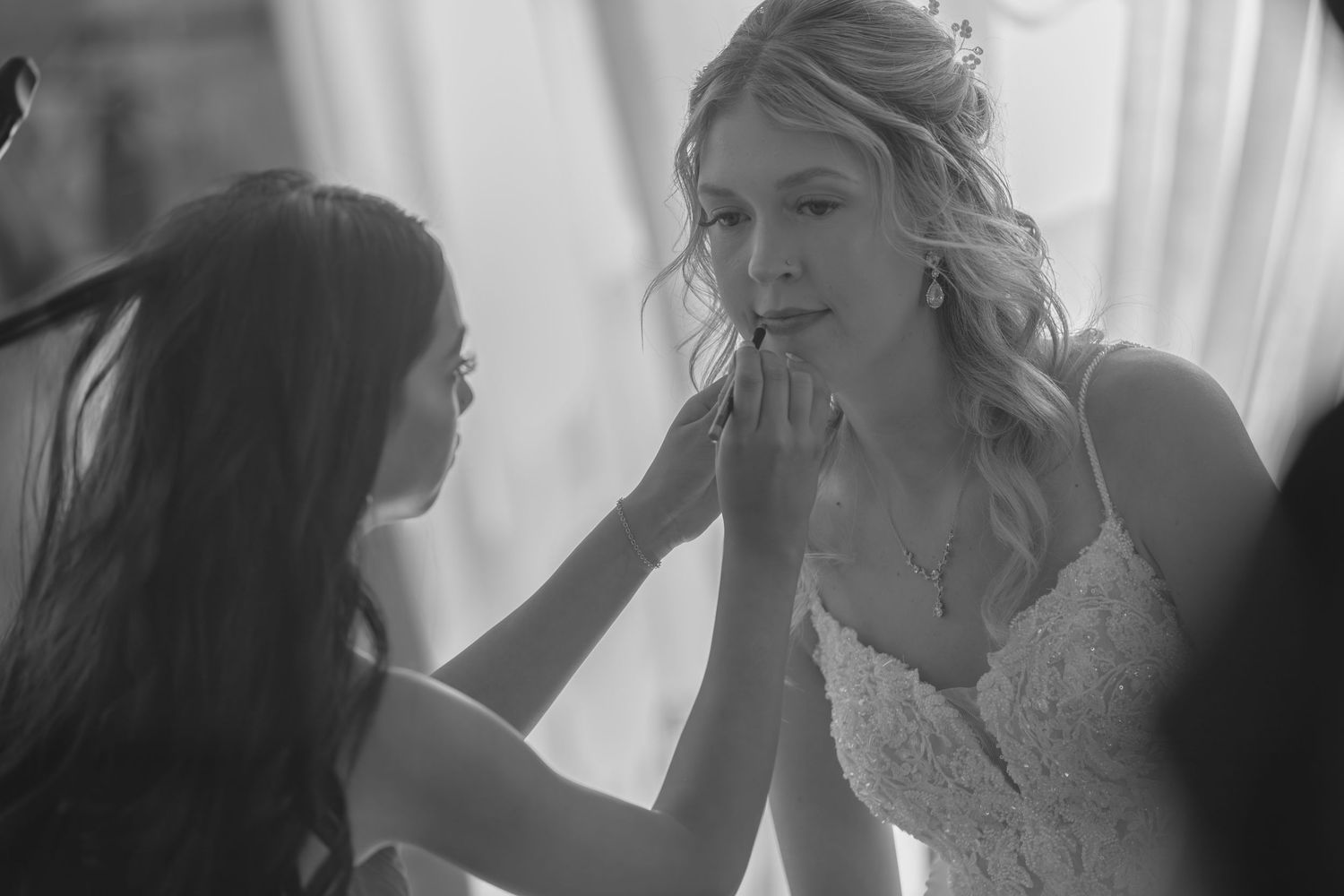A makeup artist carefully applies finishing touches to a blonde bride in a lace wedding gown before the ceremony.