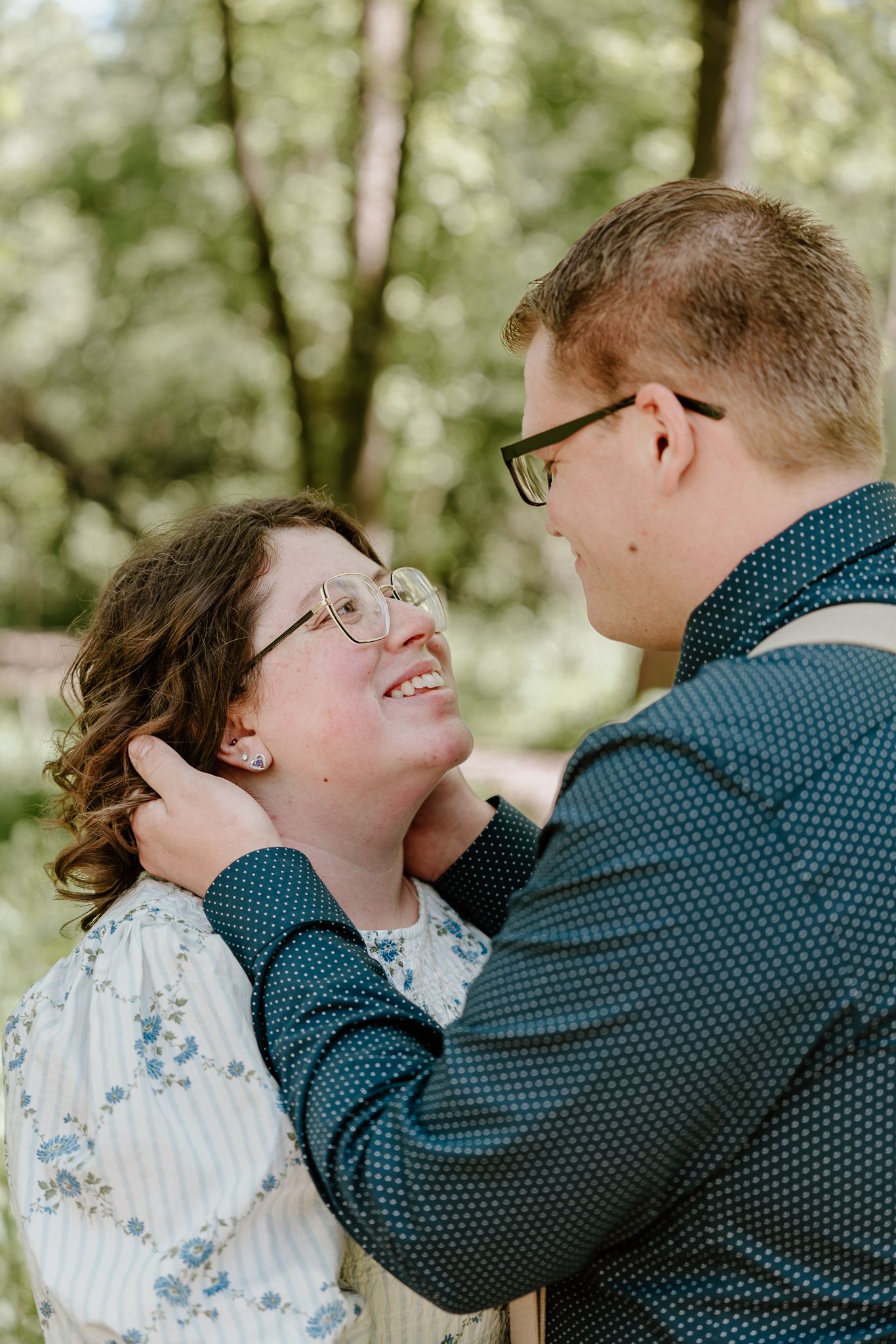 A couple shares a tender moment outdoors, man gently holding woman's face as she smiles up at him, both wearing glasses.