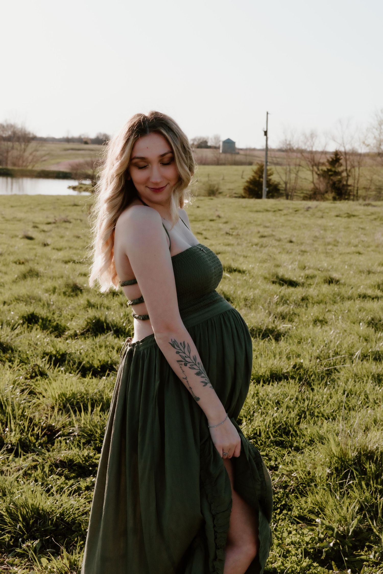 Expectant woman in a flowing green dress poses outdoors in a pastoral field near a pond at sunset.