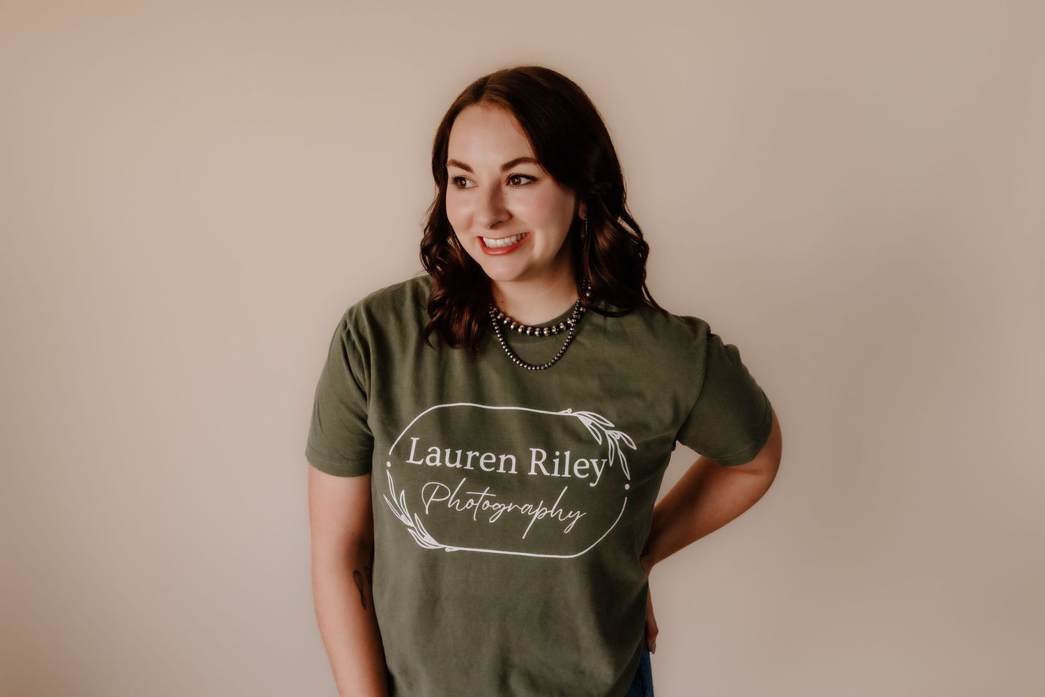 Someone wearing an olive green t-shirt with logo typography against a beige background poses for a photo.