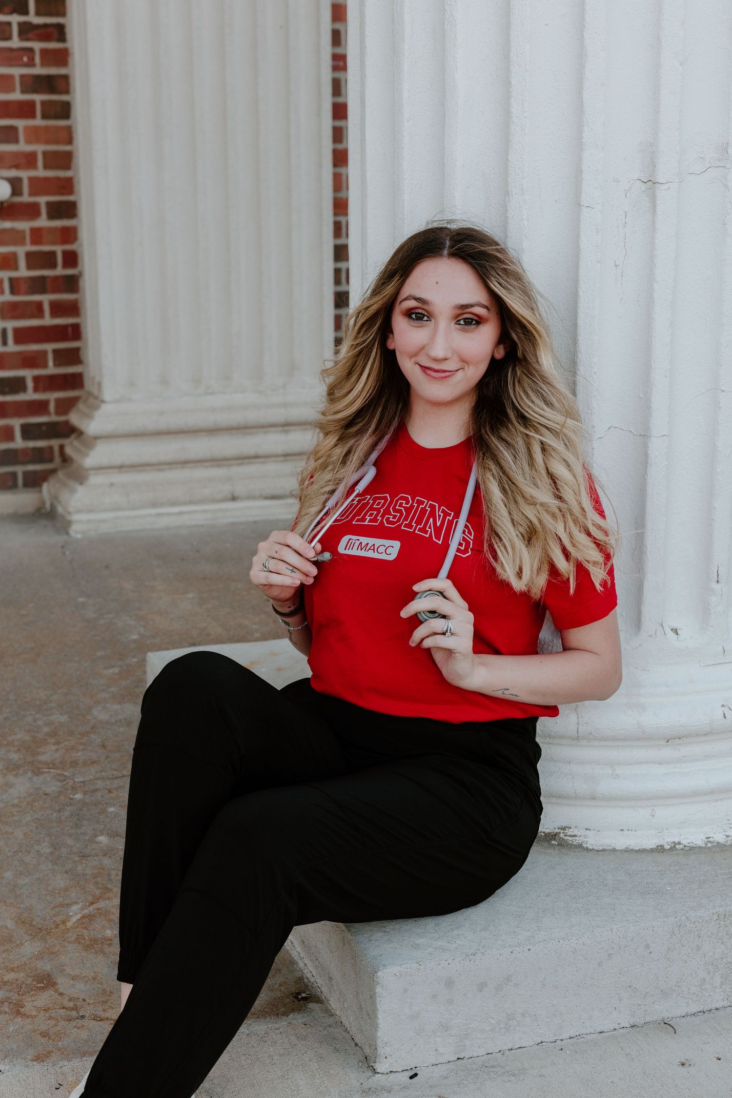 Student in red university t-shirt poses next to white outdoor column for graduation photos.