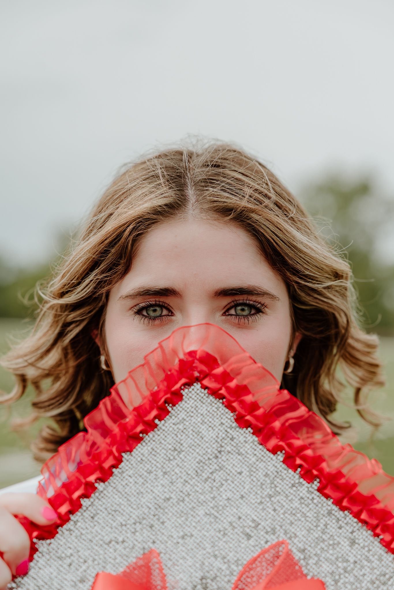 Young woman with green eyes peering over a silver and red ruffled graduation cap outdoors.