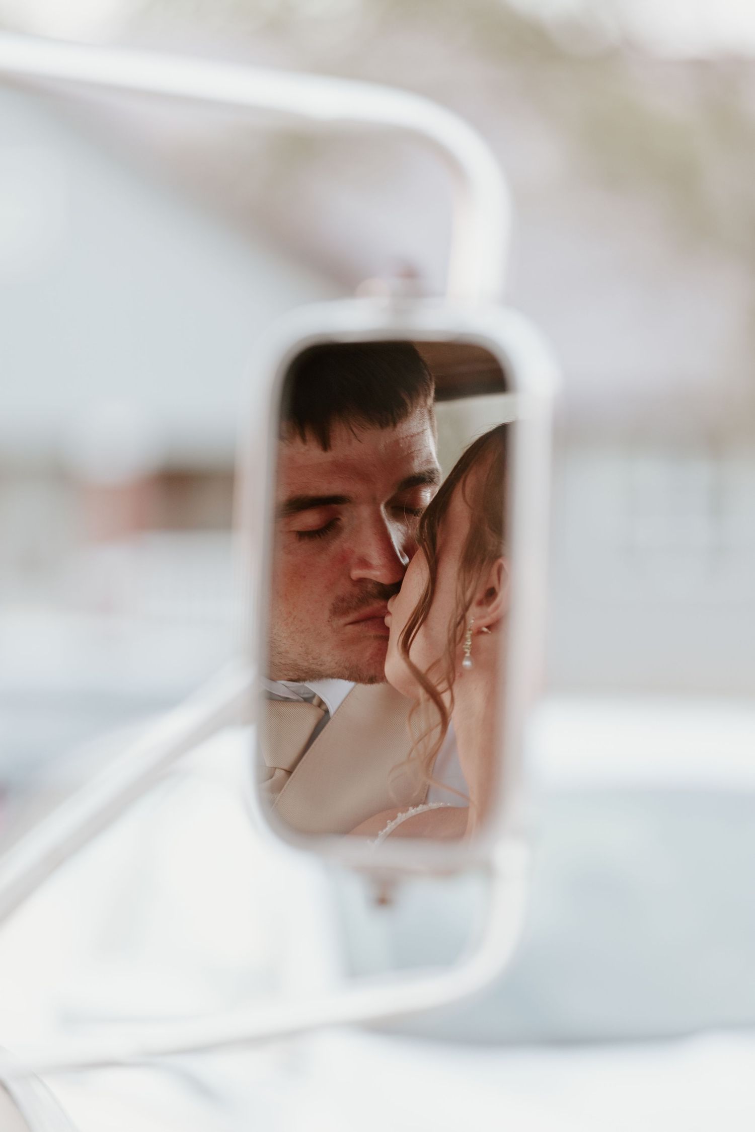 Bride and groom sharing a romantic kiss reflected in a vintage car side mirror on their wedding day.