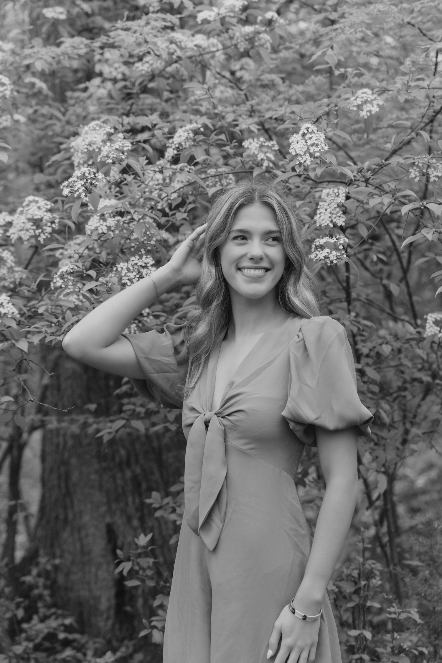 Woman in gray dress poses outdoors in front of flowering trees during a summer photoshoot.