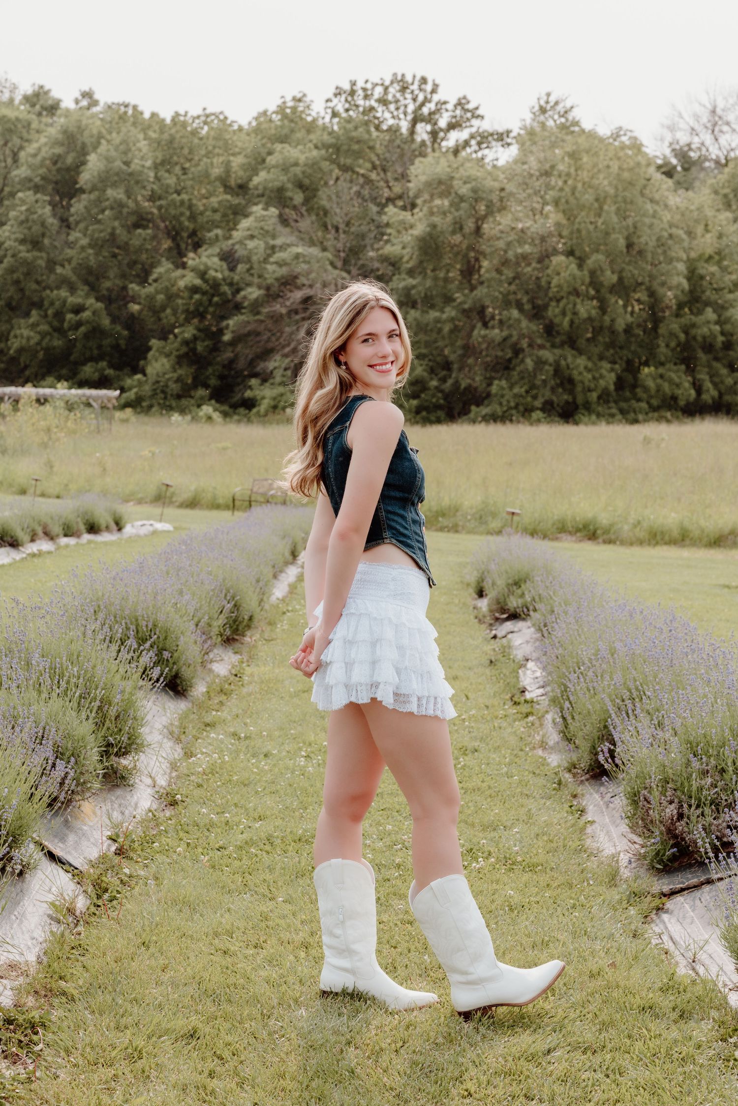 A person in white boots and a ruffled skirt poses between rows of blooming lavender plants in a sunlit garden.