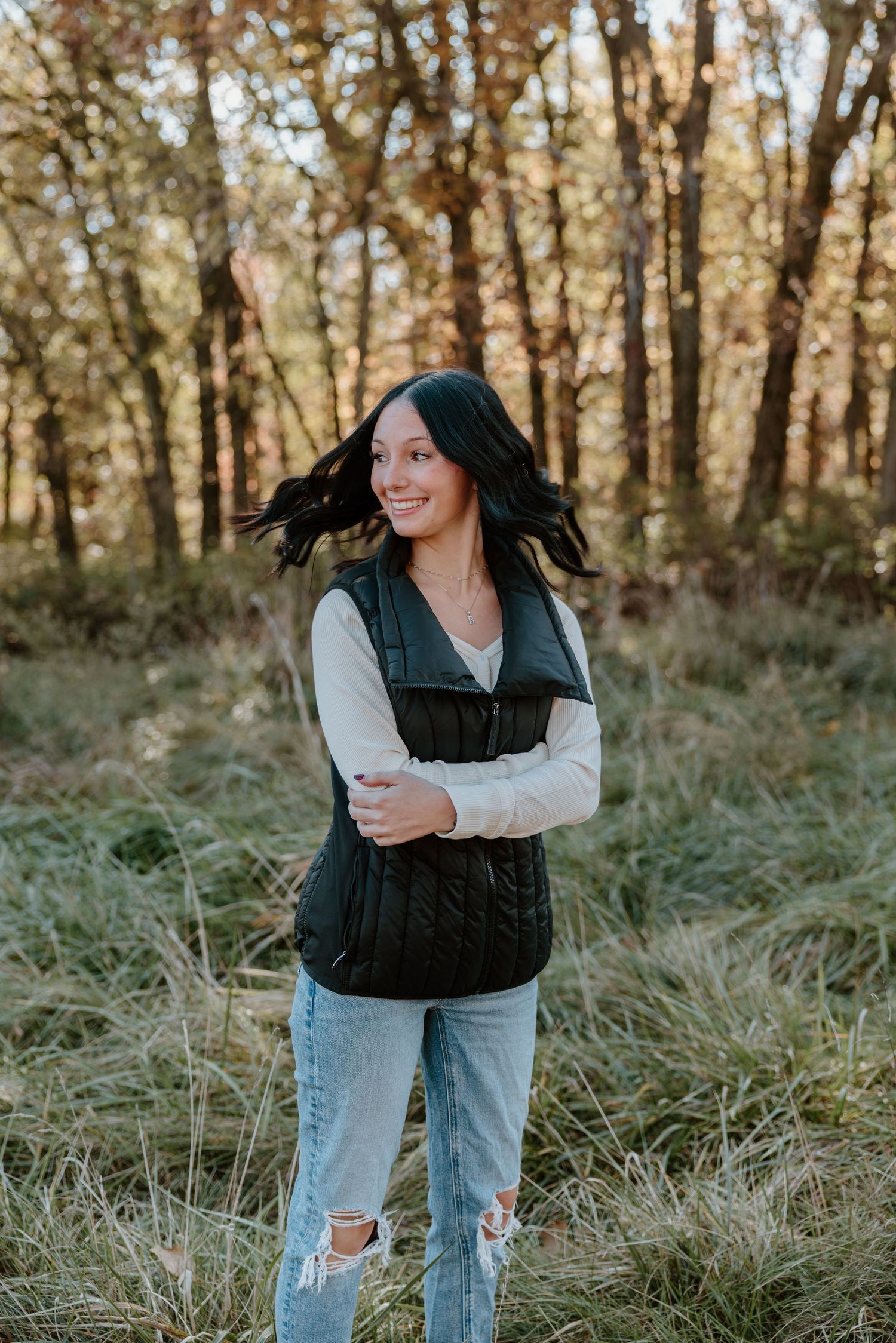Person in ripped jeans and black vest stands in tall grass during golden hour in autumn forest.