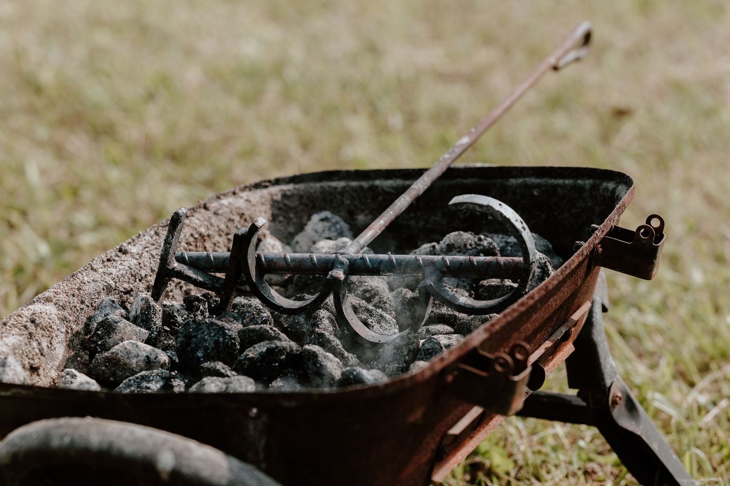 Rusty wheelbarrow filled with smoldering charcoal and metal tools on grass outdoors.