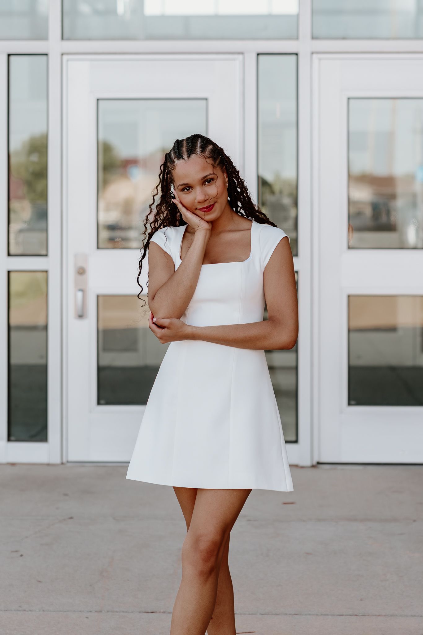 Young woman in white mini dress with braided hair posing confidently in front of glass doors.