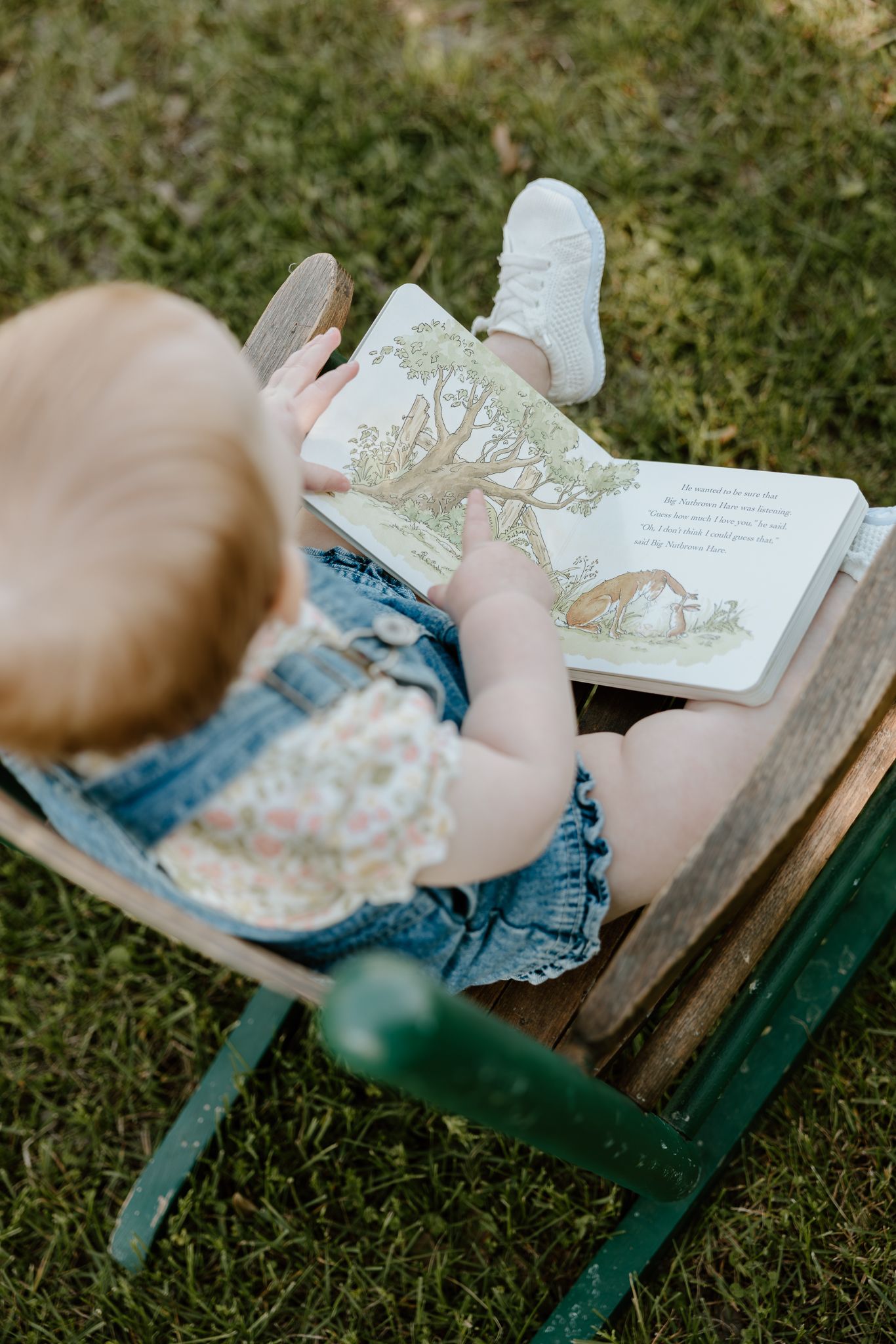 A baby girl in denim overalls sits in a wooden chair outdoors, reading a picture book on a grassy lawn.