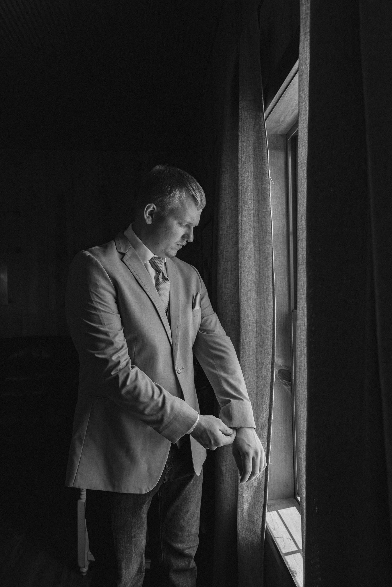 Black and white photo of a well-dressed man in a suit adjusting his cufflinks by a window in a dark room.