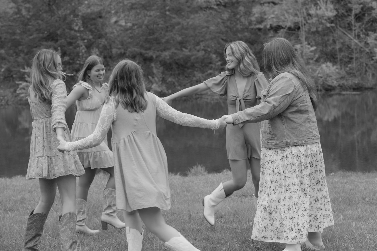 A black and white photograph shows a joyful group dancing and holding hands in a circle by a lake.