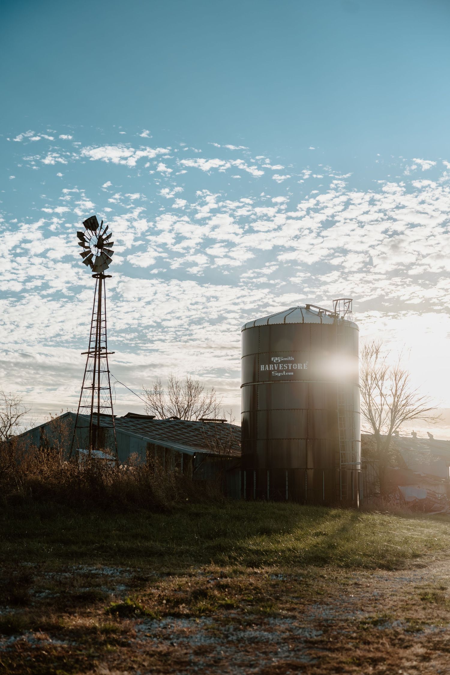 Silhouetted grain silos and windmill against a bright cloudy sky at sunset in a rural landscape.
