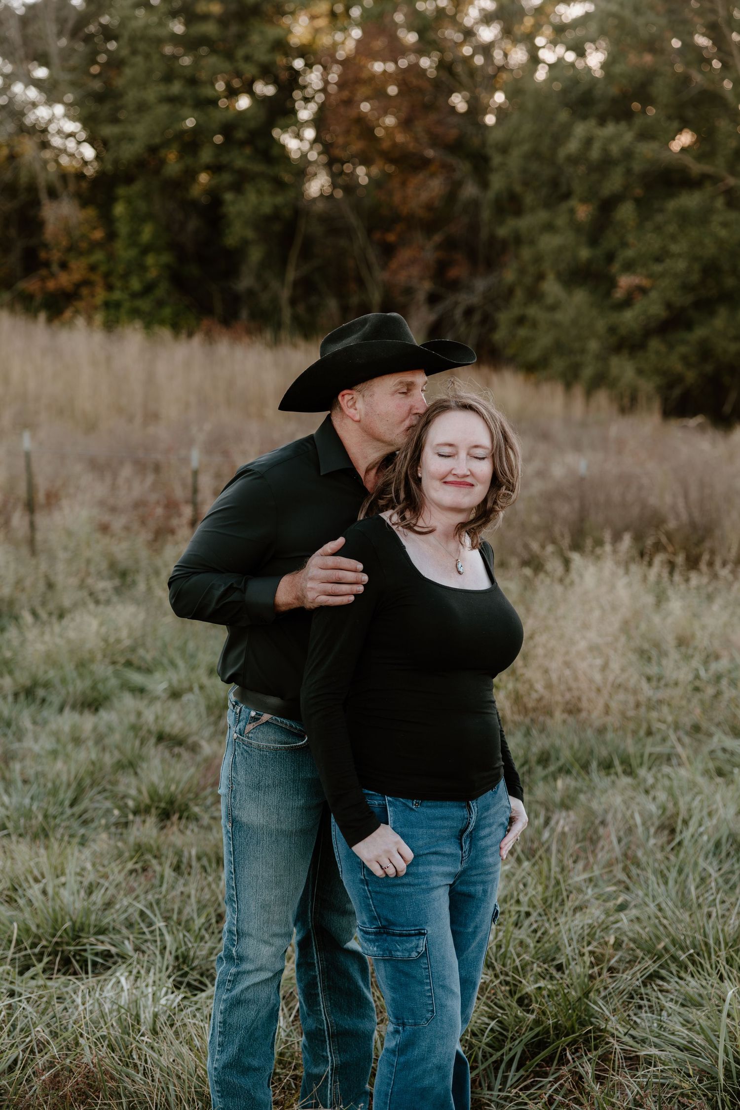 A couple in casual denim and black attire share tender moments in a grassy field during golden hour.