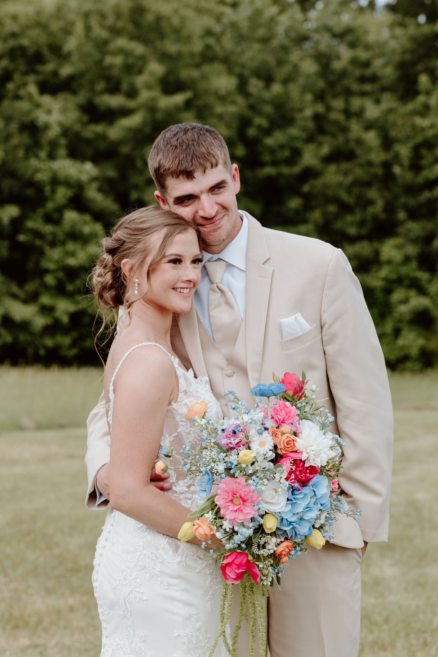 Bride and groom pose outdoors, she holds a colorful bouquet, he wears a tan suit, green trees in background.