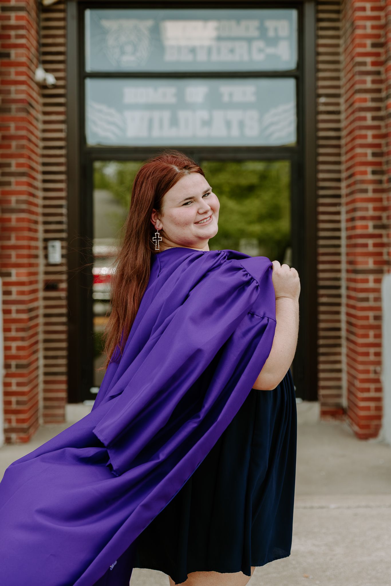 A smiling young woman in a purple graduation gown poses in front of a brick building with a Home of the Wildcats sign.