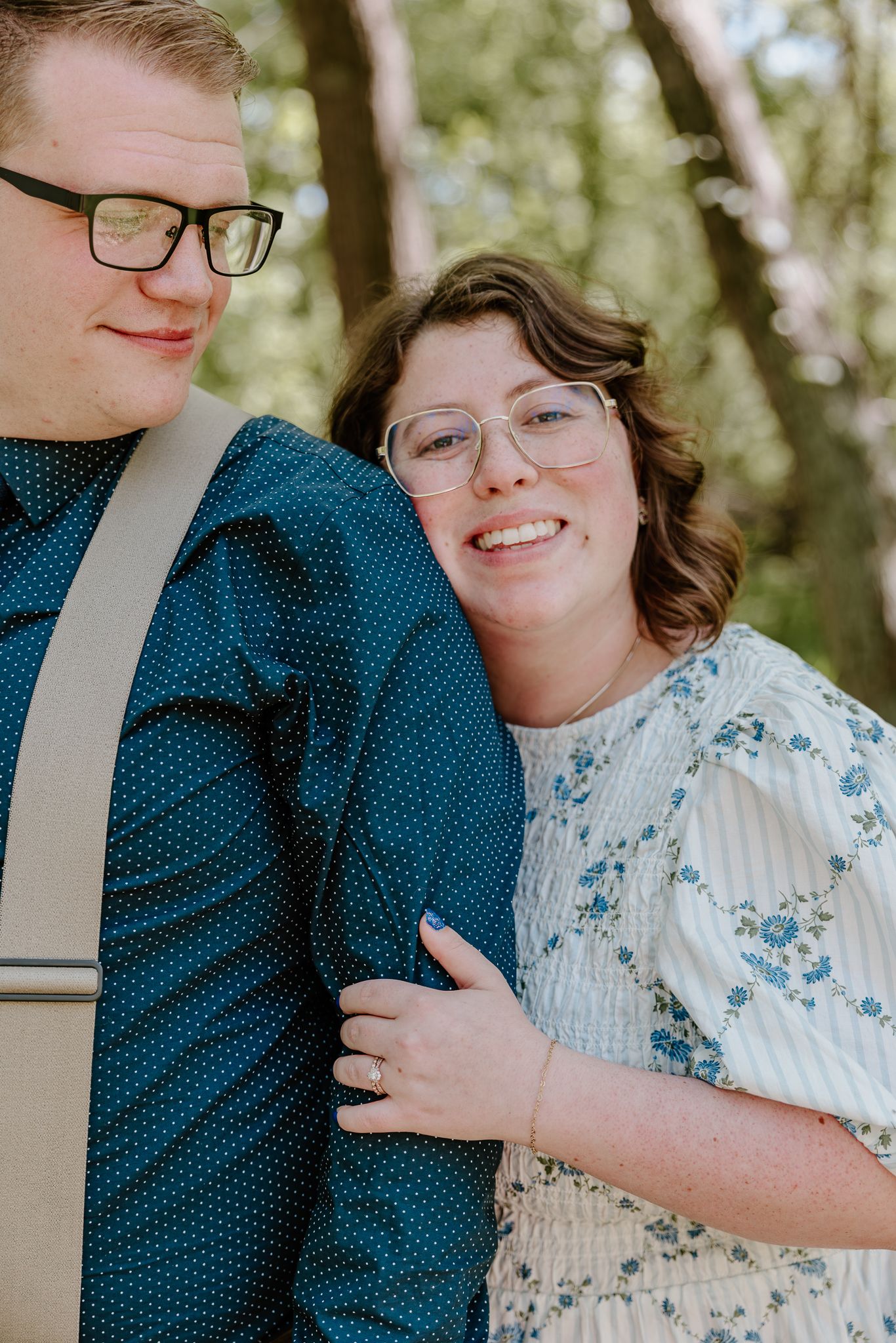 A smiling couple in glasses pose outdoors, woman in floral dress leaning on man in teal polka dot shirt.
