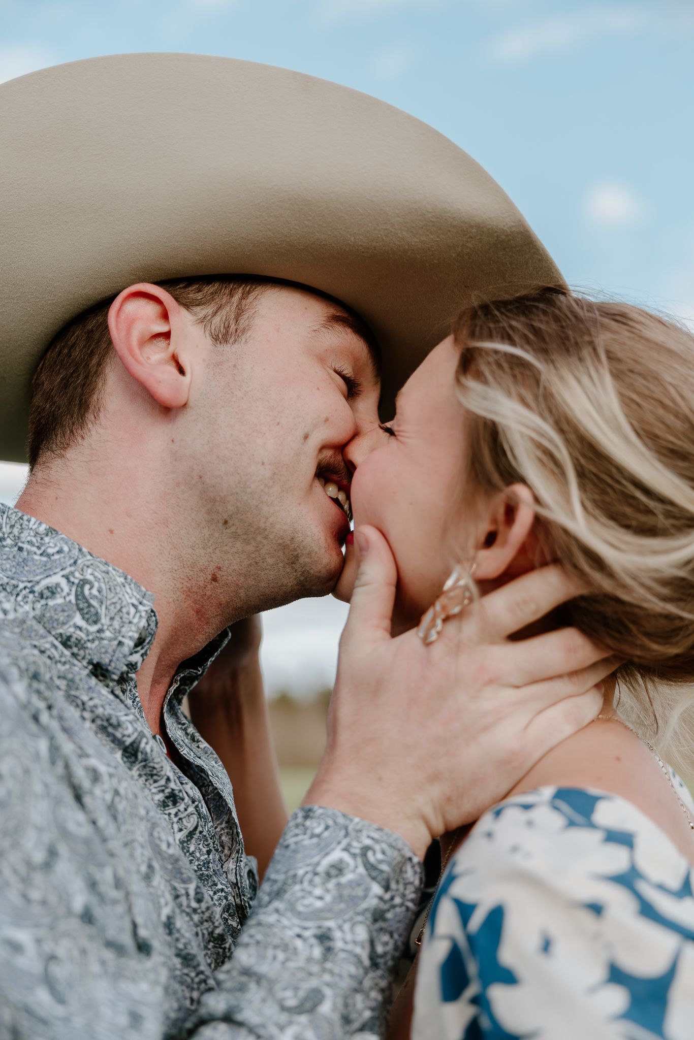 A couple shares a romantic kiss outdoors, the man wearing a cowboy hat and paisley shirt, the woman in a floral dress.