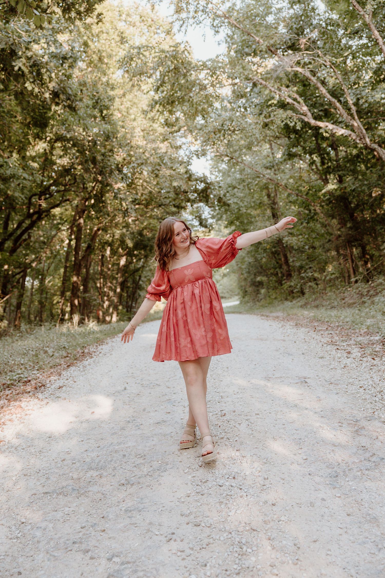 A person in a flowing red dress dances and twirls on a sunlit forest path surrounded by trees.