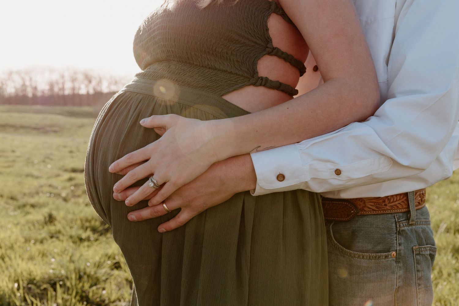 Close-up shot of a pregnant belly being embraced by loving hands in a sunlit field at sunset.